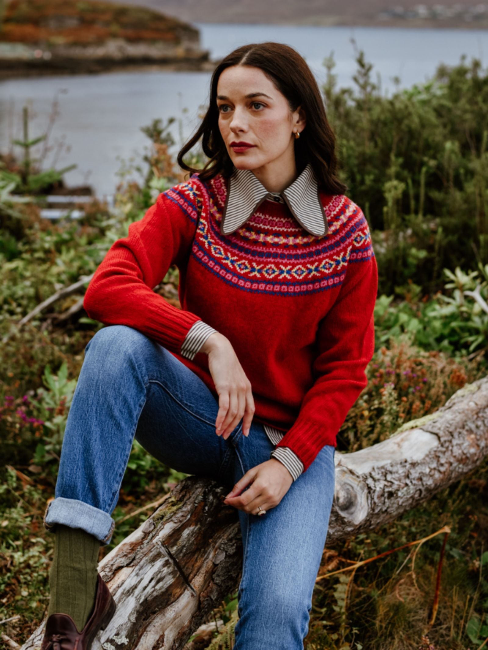 A woman in a Campbell's of Beauly Fairisle Crew Jumper and blue jeans sits on a fallen tree amid greenery near water, gazing thoughtfully into the distance.