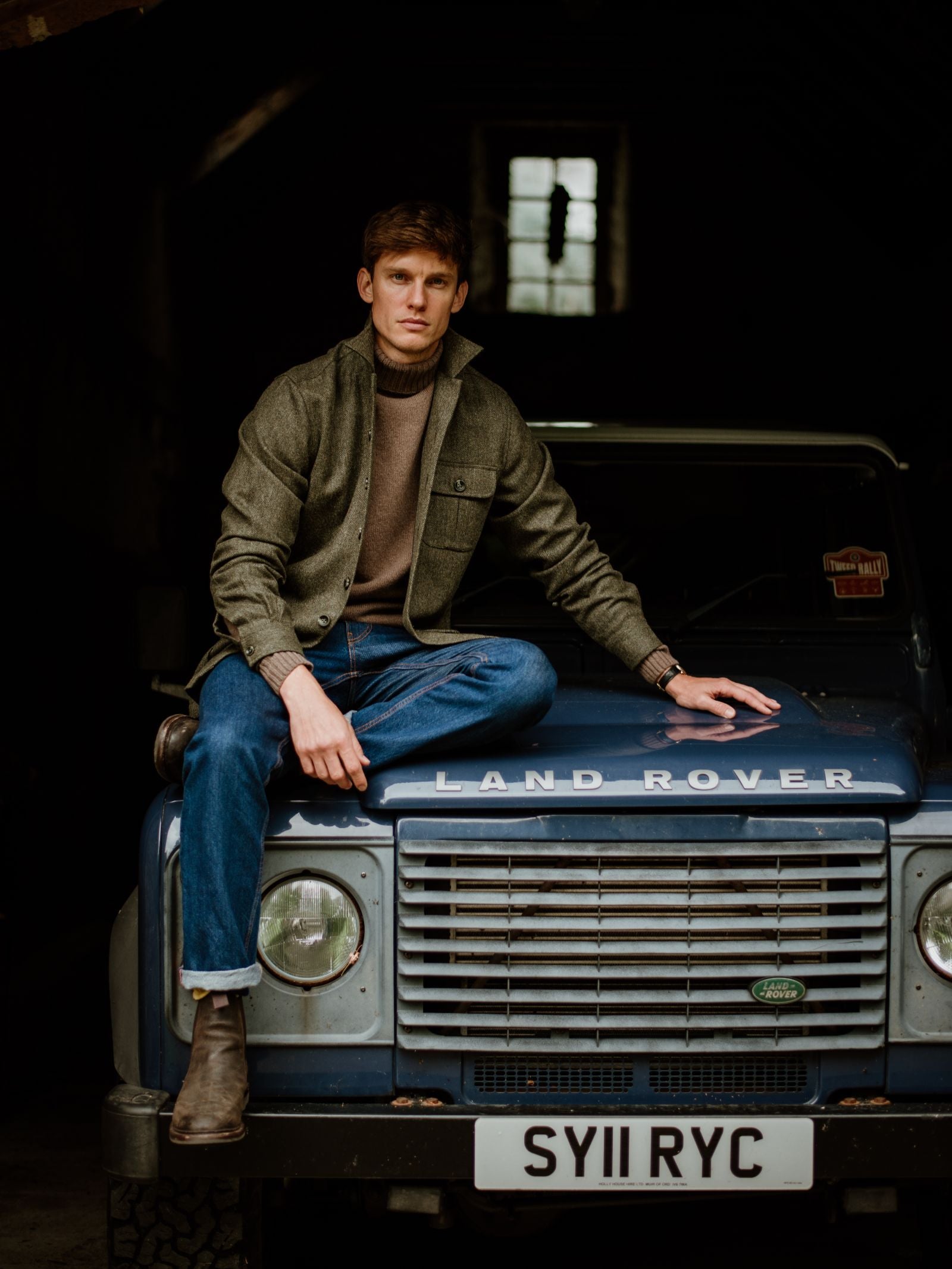 A man in a Campbell's of Beauly Casual Tweed Shacket, brown sweater, blue jeans, and boots sits on the hood of a blue Land Rover Defender (plate: SY11 RYC) inside a dimly lit garage, embodying relaxed tailoring.