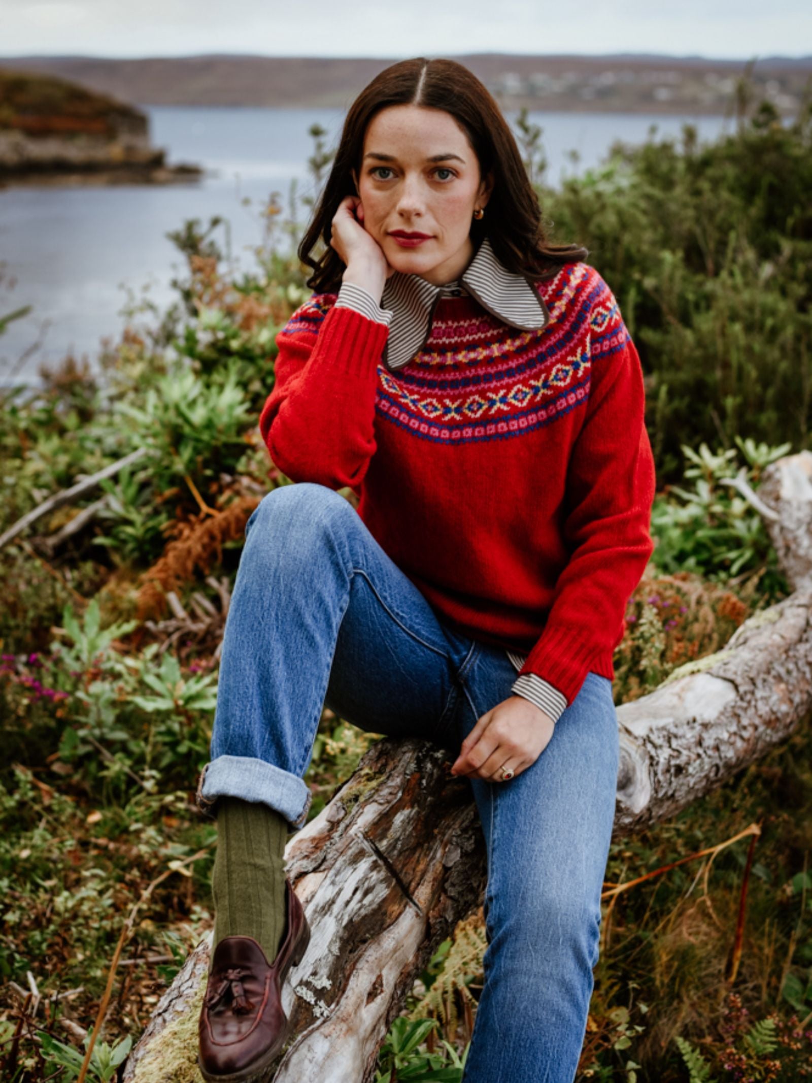 A woman in a red patterned Campbell’s of Beauly Fairisle Crew Jumper, knitted in Scotland from Geelong lambswool, sits on a fallen tree among greenery with water and hills behind her, resting her head on her hand and gazing thoughtfully at the camera.