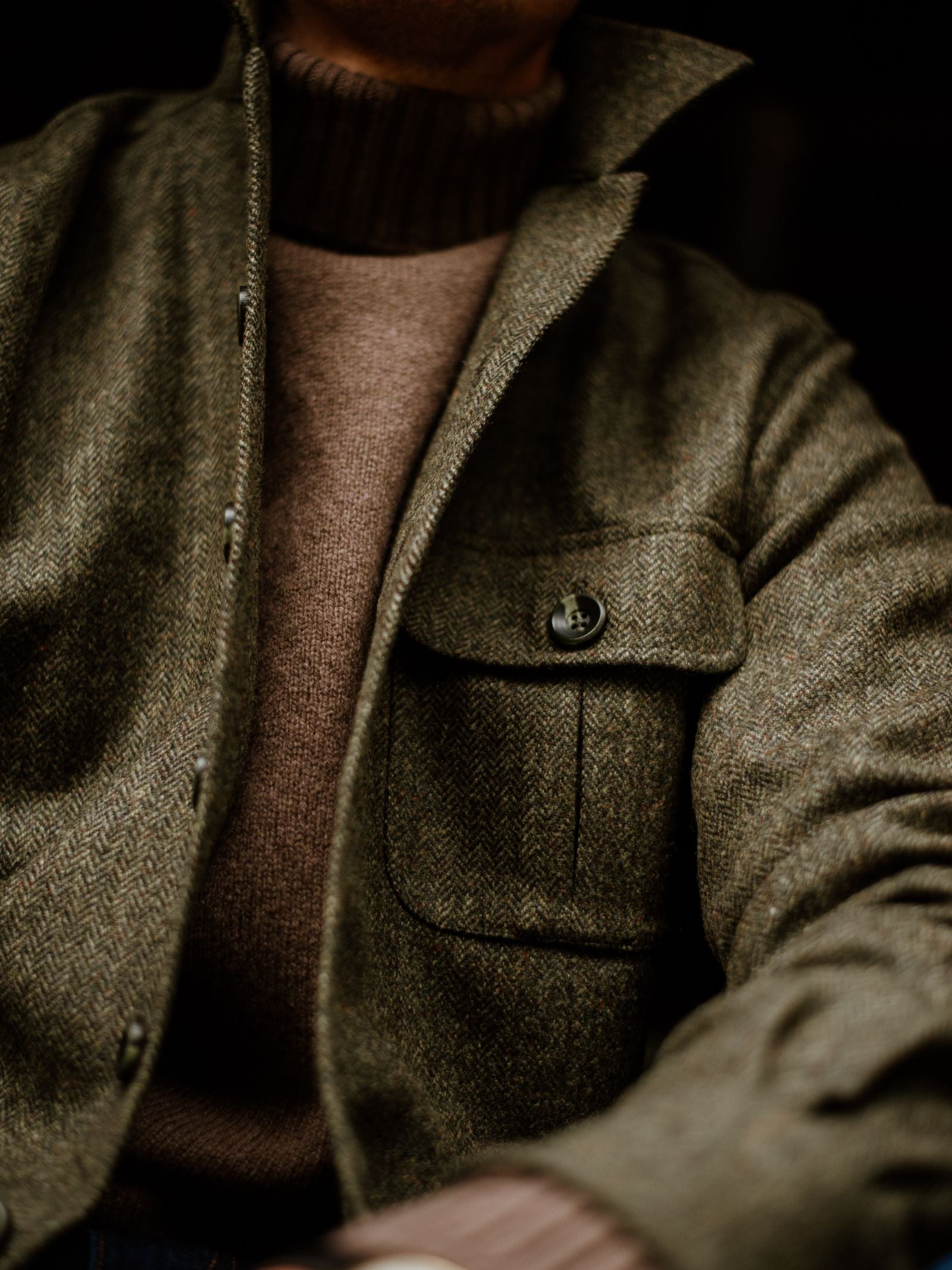 A close-up shows a person wearing the Campbell's of Beauly Casual Tweed Shacket in olive green over a brown wool turtleneck, featuring a front flap pocket and relaxed tailoring. The person's face is partially out of frame.