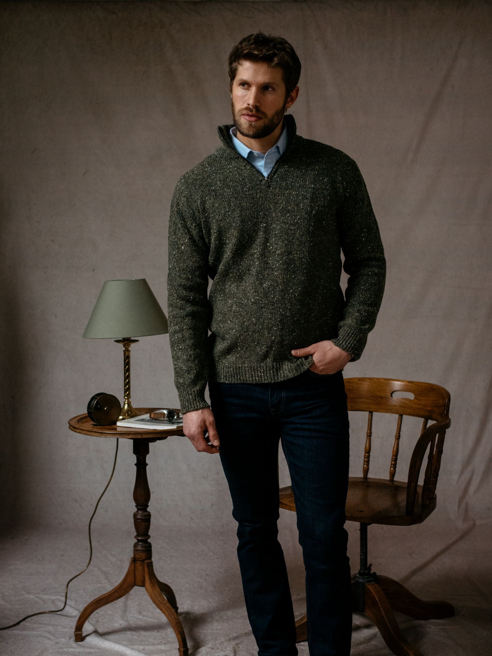 A man with short brown hair and a beard stands indoors beside a vintage wooden chair and table with a green lamp, wearing the Campbells of Beauly Donegal Lambswool Zip Neck Jumper over a collared shirt and dark jeans for an elevated country look.