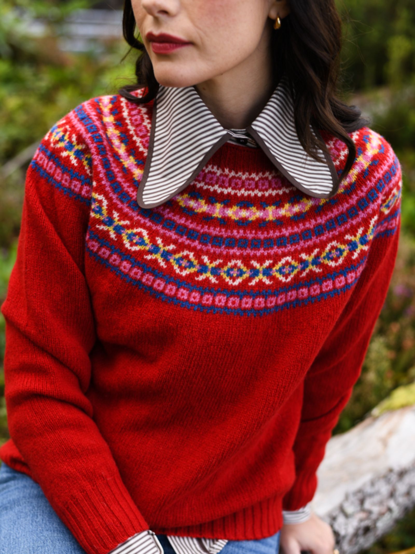 A woman wears a vibrant red Campbell's of Beauly Fairisle Crew Jumper, crafted in Scotland from Geelong lambswool, with a striped collared shirt and gold hoop earrings, as she sits outdoors among lush greenery.