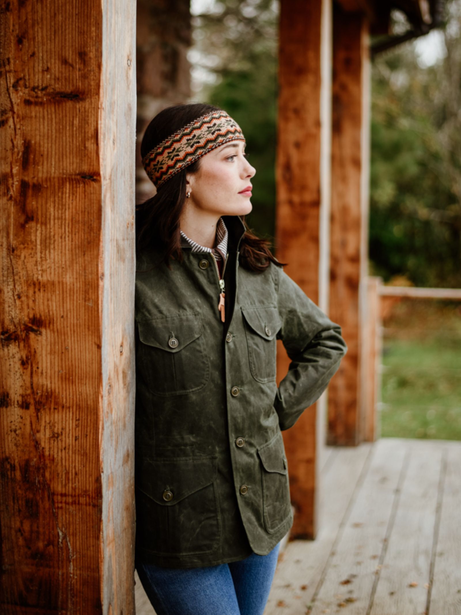 A woman in a green jacket and blue jeans wears the Campbell's of Beauly Fairisle Headband, crafted from 100% wool in the Scottish Borders, as she leans on a porch post, gazing thoughtfully with blurred trees behind her.