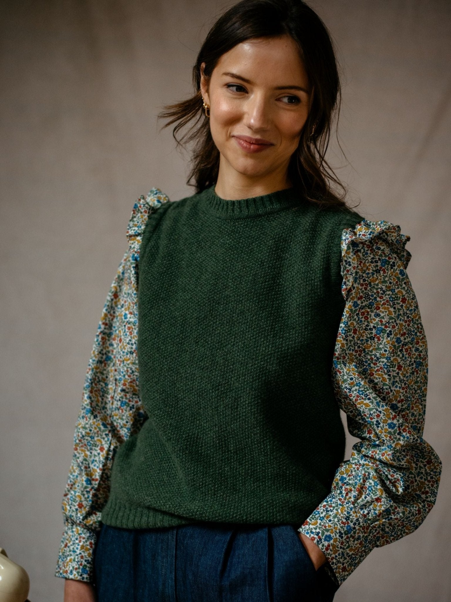 A dark-haired woman smiles gently, wearing the Campbell's of Beauly Liberty Print Peplum Shirt with ruffled shoulders under a green sleeveless sweater and dark pants, set against a neutral background.