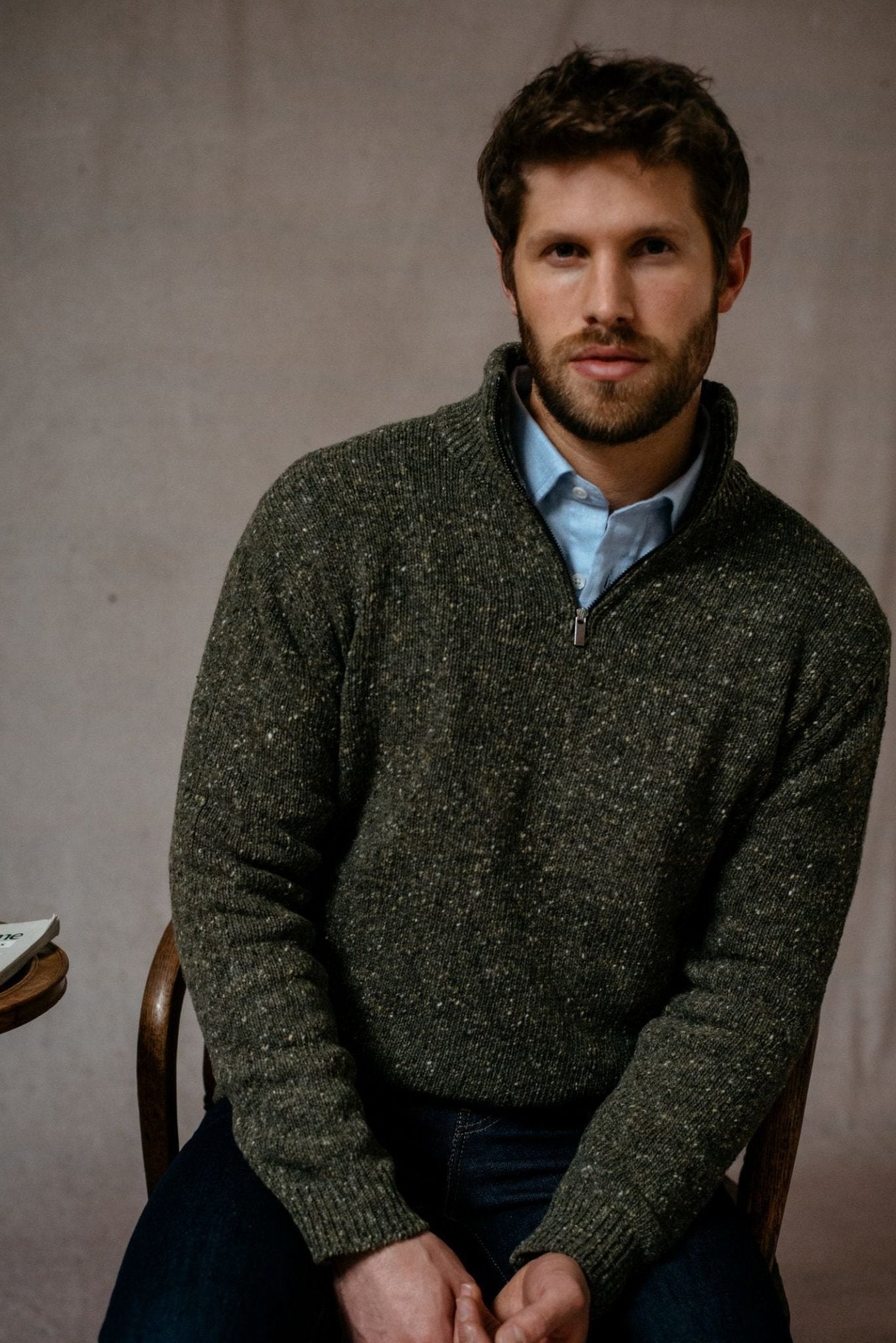 A man with short brown hair and a beard sits on a chair, wearing the Campbells of Beauly Donegal Lambswool Zip Neck Jumper over a light blue collared shirt—a timeless addition to any country wardrobe.