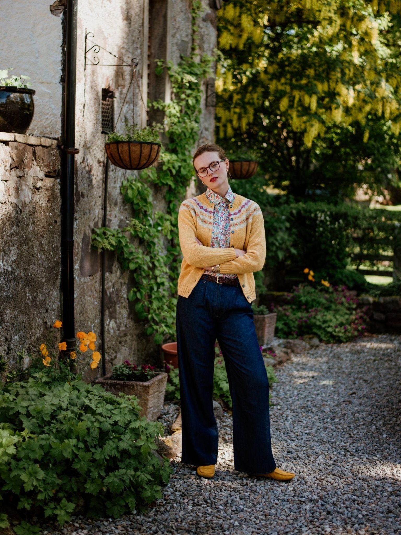 A person with short hair, glasses, and red lipstick stands with arms crossed outdoors, wearing a yellow cardigan, patterned shirt, Campbells of Beauly Wide Leg Jeans in lightweight denim, and yellow shoes near greenery by a stone wall.
