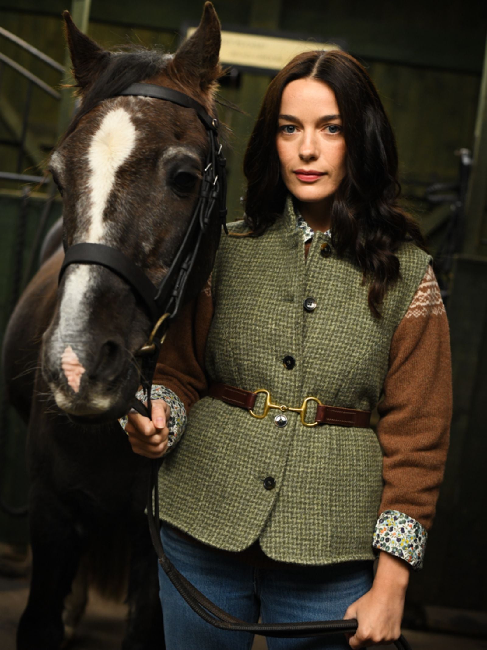 A woman with long dark hair wears the Campbell's of Beauly Auld Stock Harris Tweed Houndstooth Gilet as she stands in a stable, holding the bridle of a black horse with a white facial stripe.