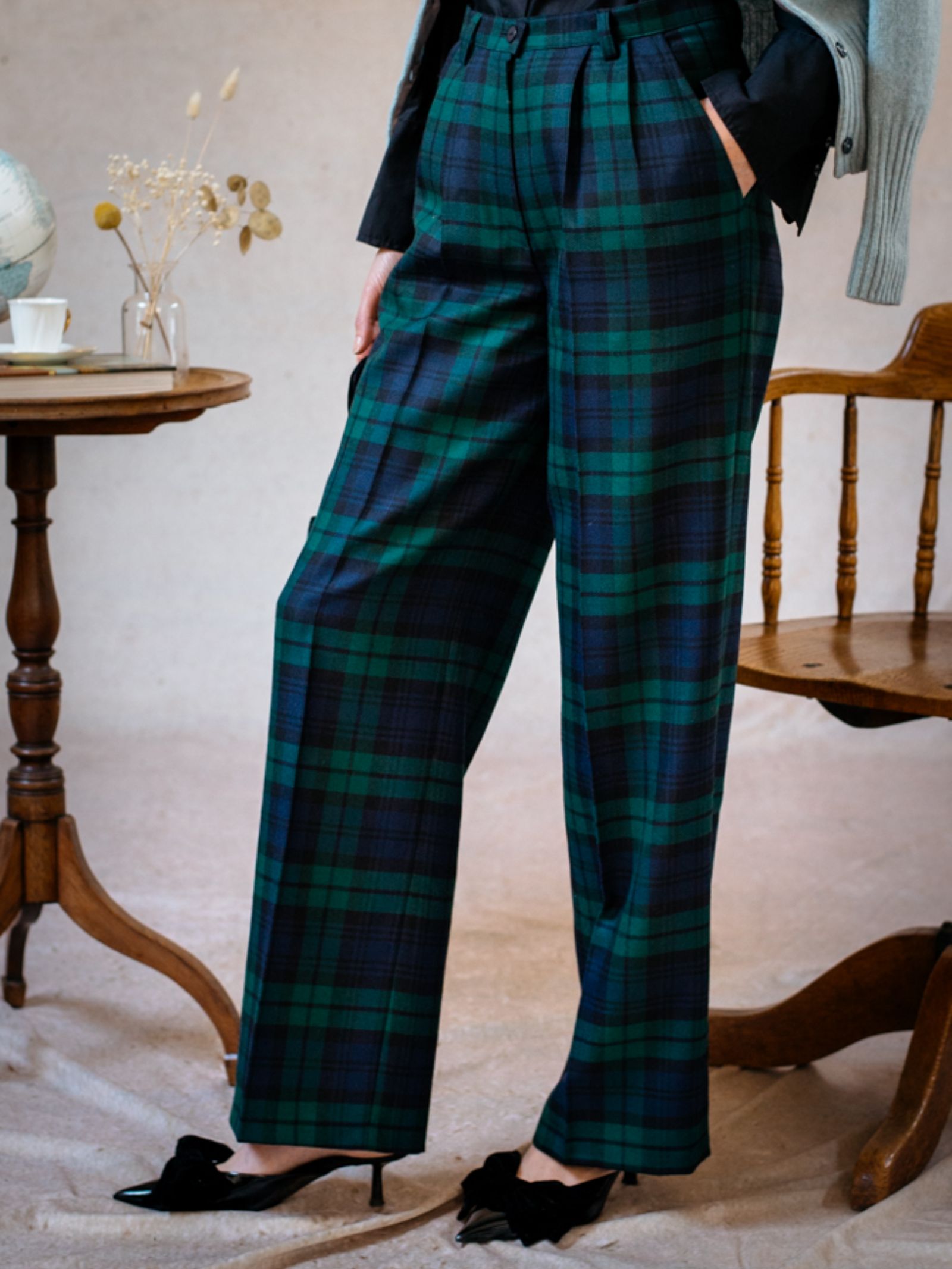 A person models the Campbells of Beauly Wide Leg Blackwatch Trousers, showing only their lower half indoors with black heels. A wooden chair and a small table with a teacup and dried flowers are next to them.