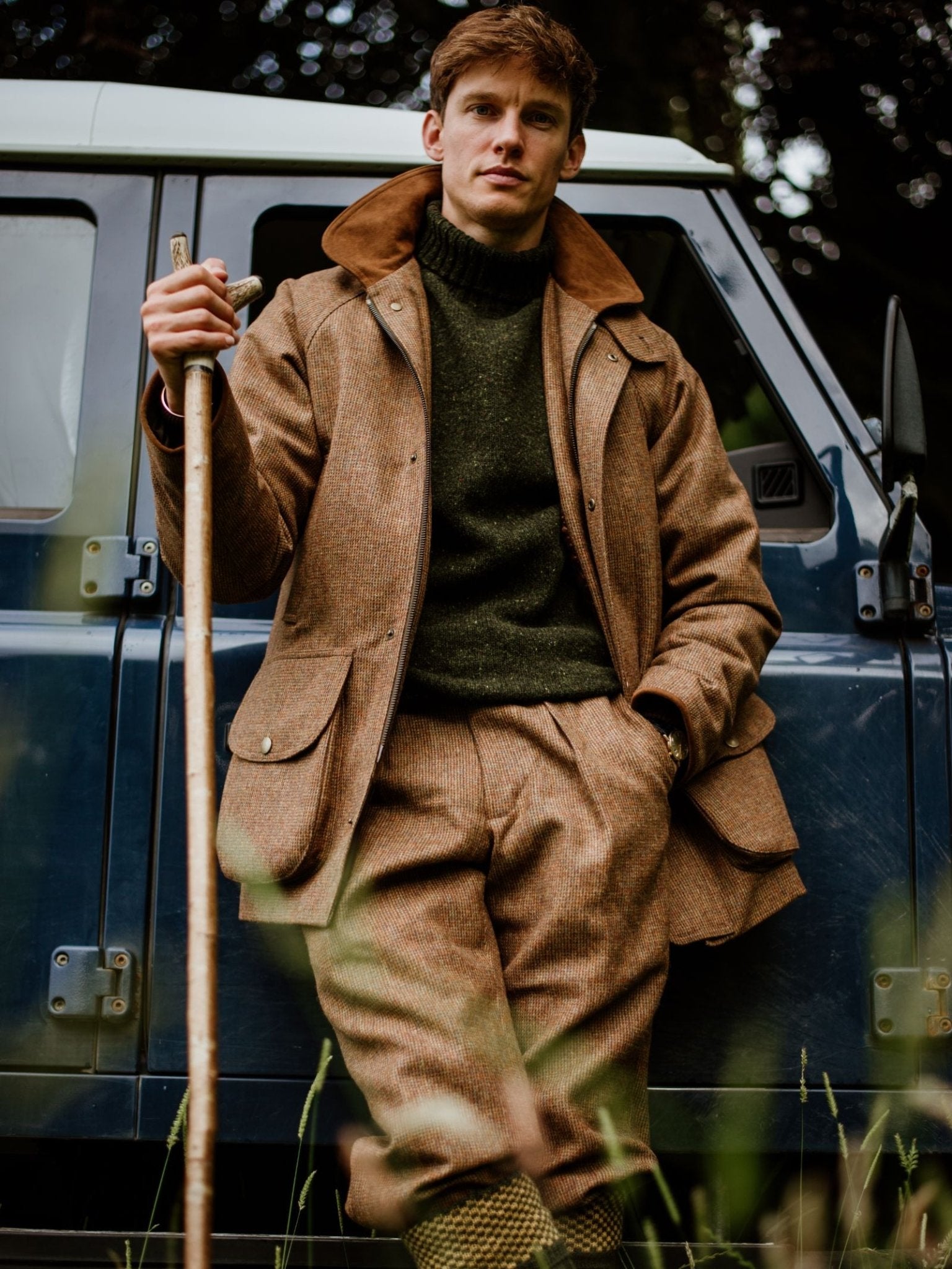 A man in tweed outdoor clothing styled for the countryside stands by a blue off-road vehicle, wearing Campbells of Beauly Tweed Shooting Waistcoat—a classic piece perfect for pairing with a field gilet. Grass and trees frame the scene.