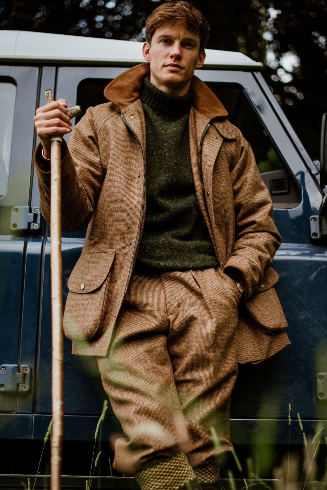 A man wearing Campbells of Beauly Tweed Breeks, a green sweater, and knee-high socks leans against a blue off-road vehicle with a wooden walking stick, dressed in classic country style amid trees.