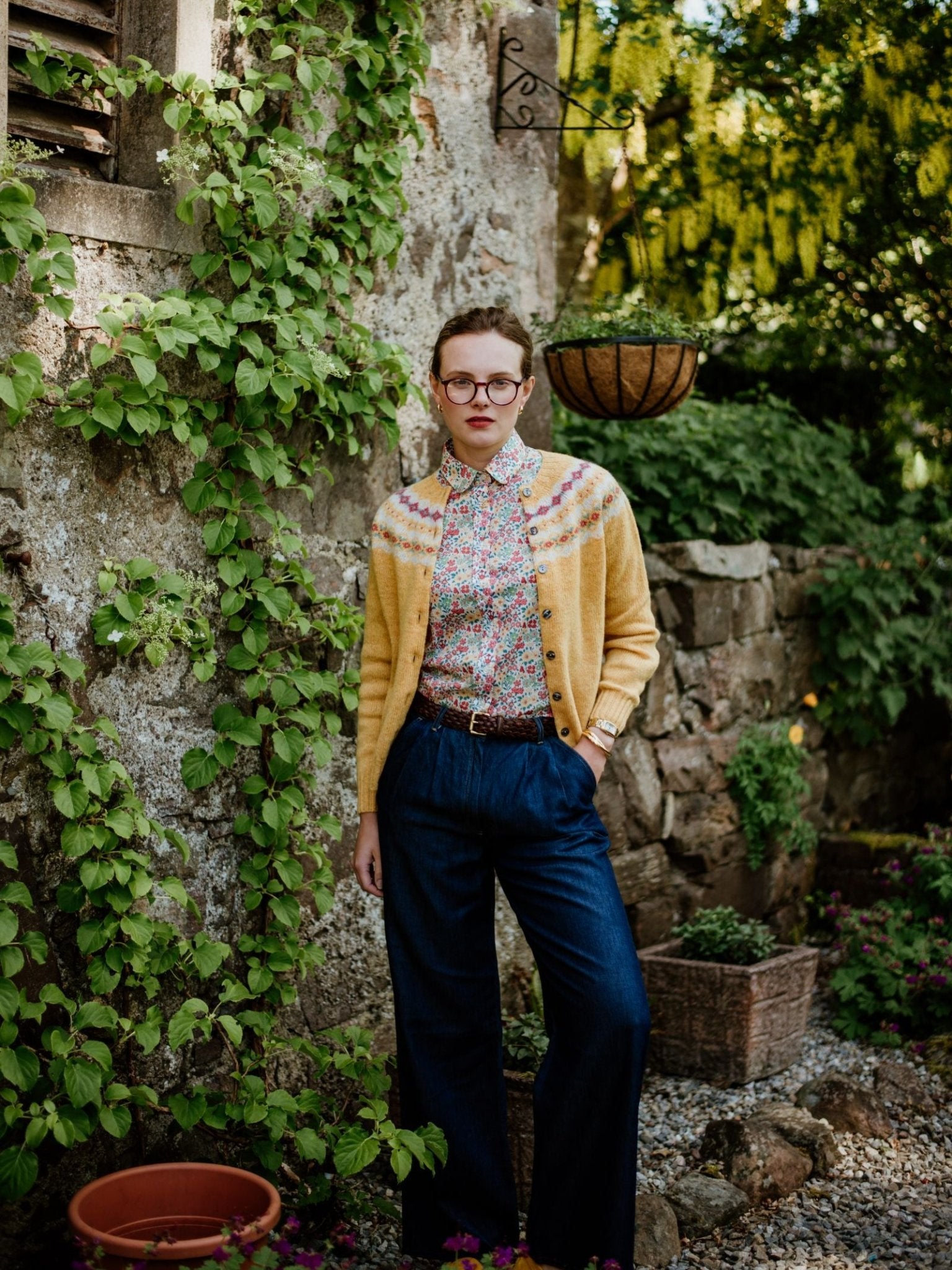 A person wearing glasses, a Campbells of Beauly Shetland Fairisle Cardigan, a floral blouse, and blue high-waisted pants stands in a lush garden by an old stone wall covered in green vines.