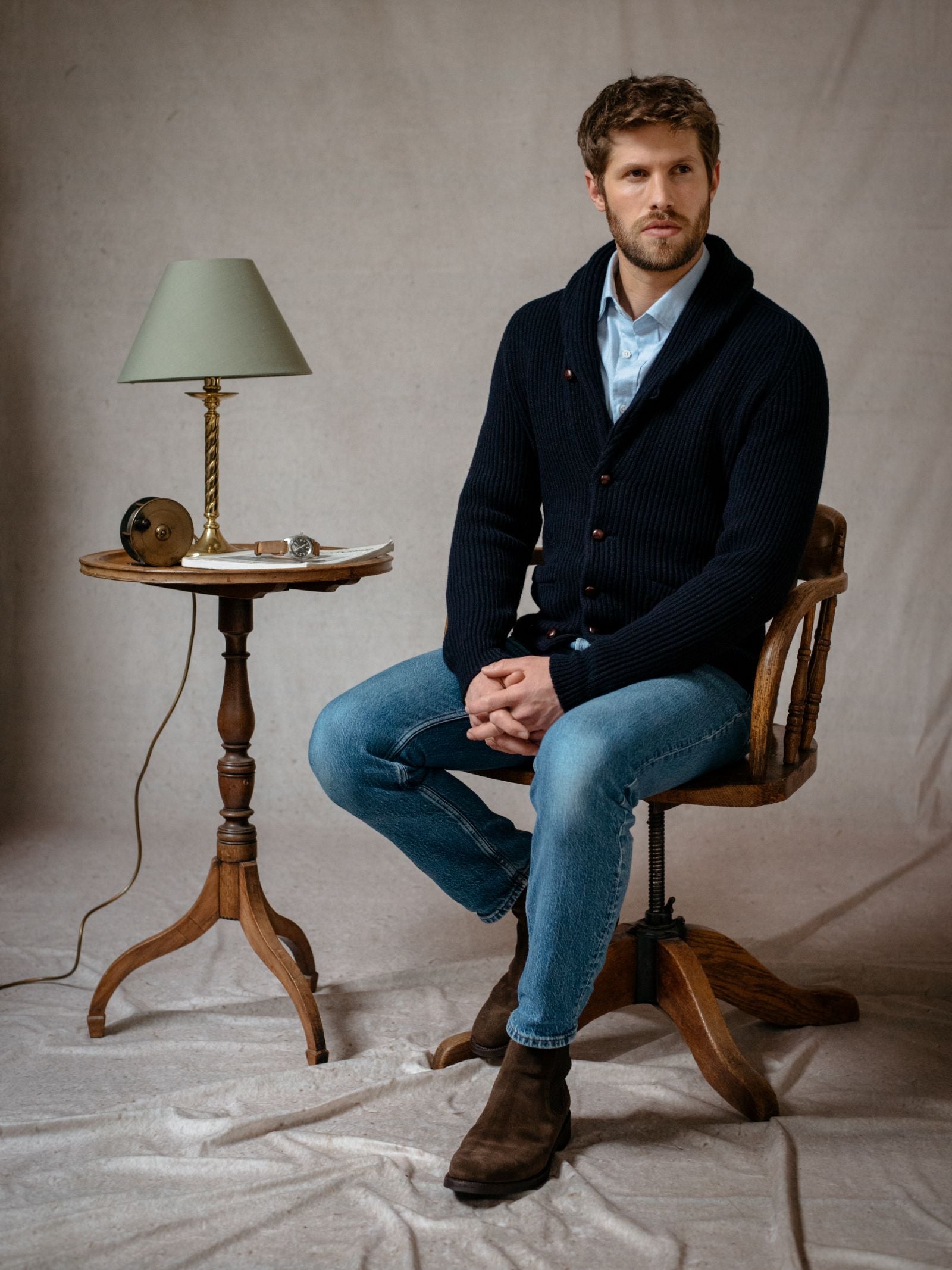 A man with light brown hair and a beard wears the Campbells of Beauly Shawl Collar Cardigan, navy, over a light blue shirt, jeans, and brown boots as he sits on a wooden chair by a table with a lamp, book, pen, and reel against a neutral backdrop.