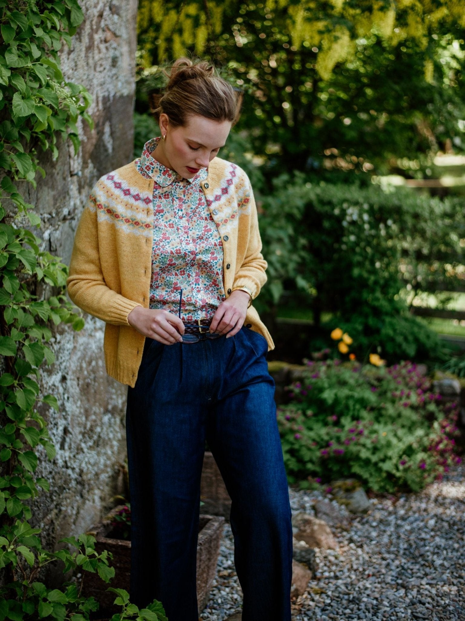 A woman stands by a stone wall in a garden, wearing a yellow Campbells of Beauly Shetland Fairisle Cardigan, a floral shirt, and high-waisted dark blue trousers as she adjusts her waistband among lush greenery.