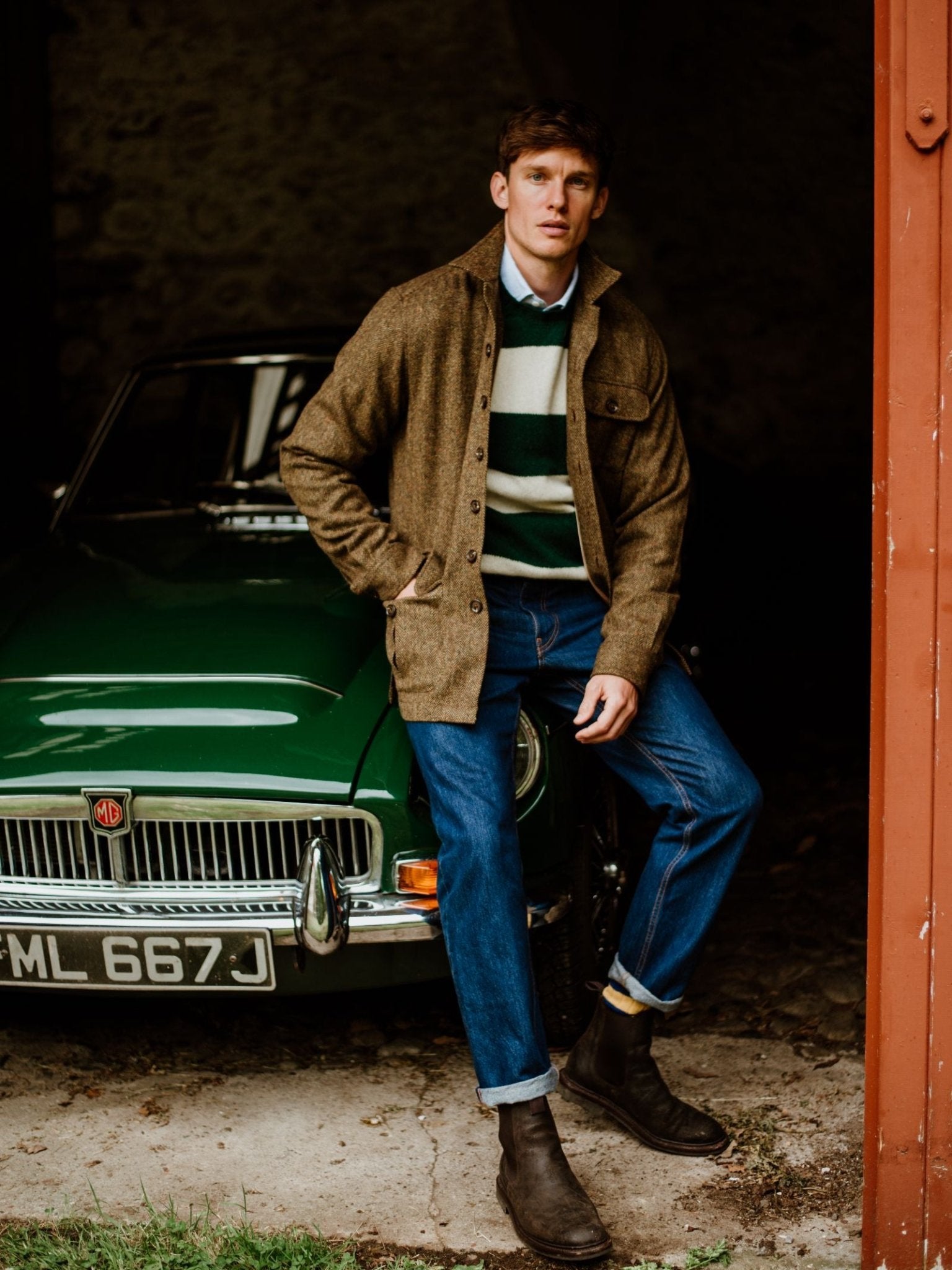 A man in a Campbell's of Beauly Casual Tweed Shacket, green and white striped sweater, blue jeans, and brown boots sits on the front of a classic green MG car inside a garage.
