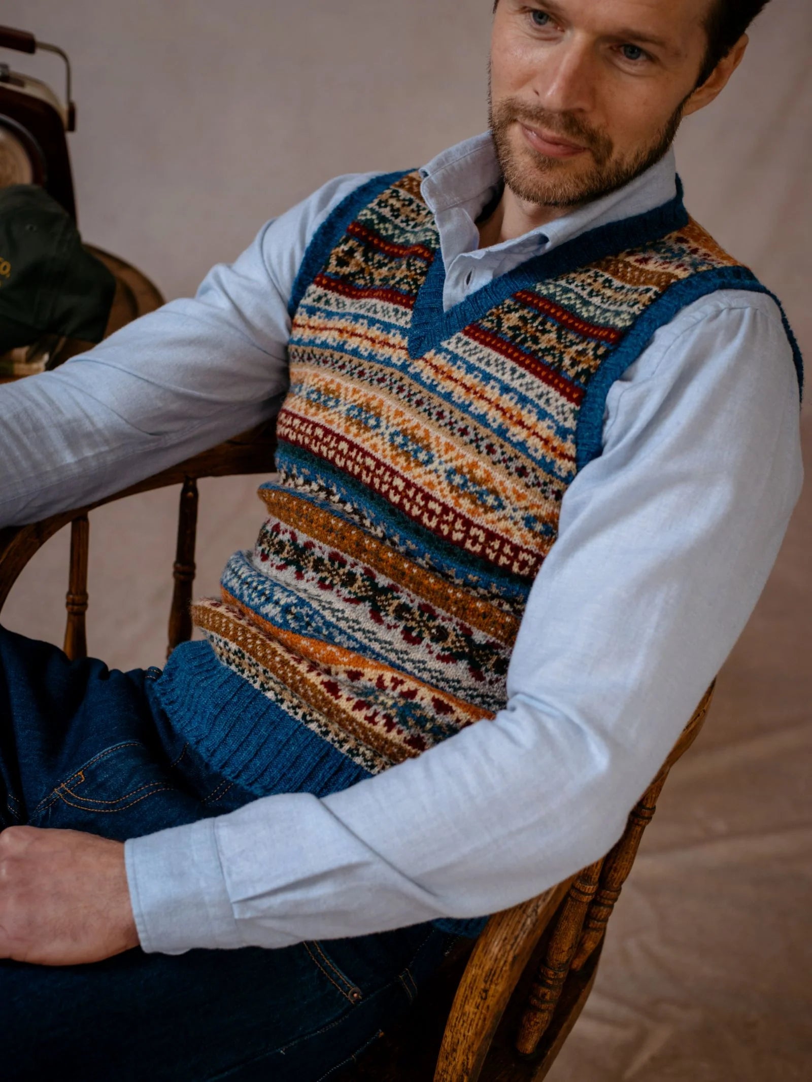 A man sits on a wooden chair wearing a light blue shirt and the Campbell’s of Beauly Fairisle Slipover—a sleeveless layer crafted from Shetland wool, featuring blue, gold, red, and white designs. He has short hair and a trimmed beard, gazing slightly off to the side.