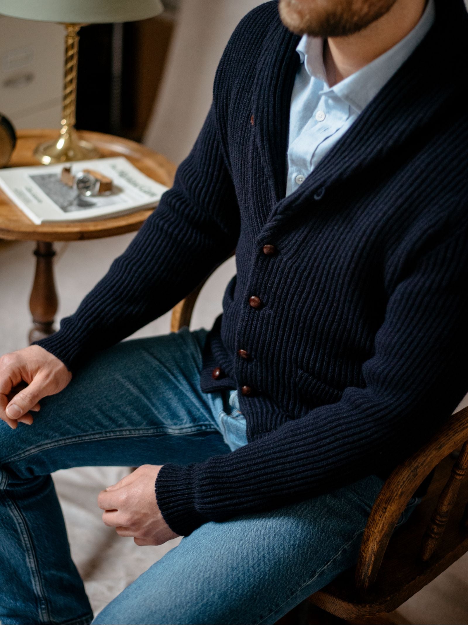 A man in a Campbells of Beauly Shawl Collar Cardigan, light blue shirt, and blue jeans sits on a wooden chair next to a round table with books, glasses, and a green lamp. His face is partially out of frame.
