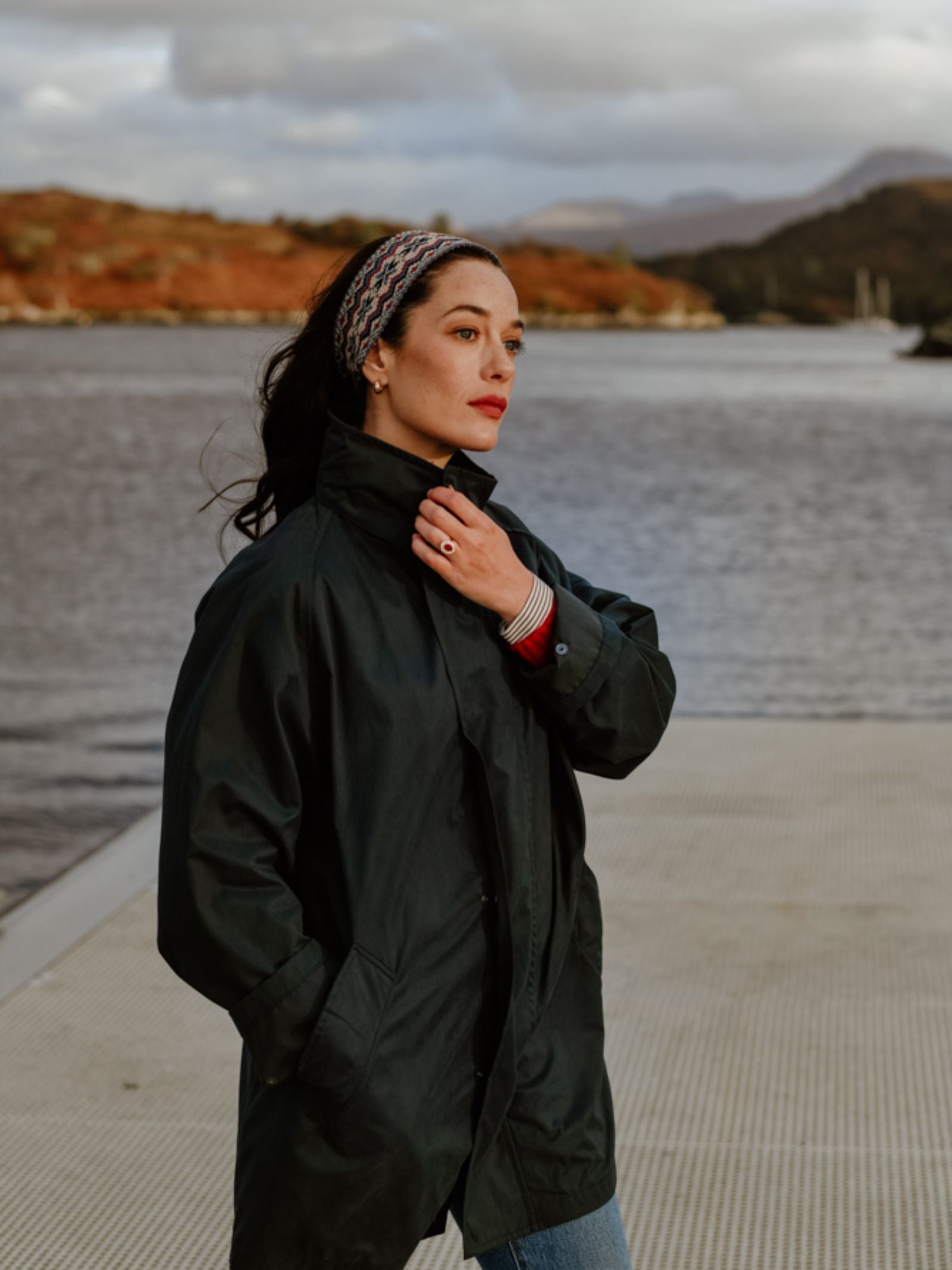 Wearing a Campbell's of Beauly Fairisle Headband and a dark coat, a woman stands on a dock by the water, gazing thoughtfully at the autumn hills and cloudy skies that capture the serene spirit of the Scottish Borders.