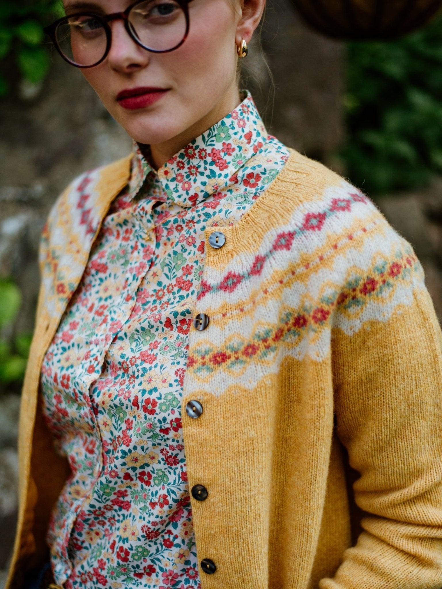 A woman with glasses and red lipstick wears a Campbells of Beauly Shetland Fairisle Cardigan over a floral button-up shirt, standing outdoors with greenery in the background.