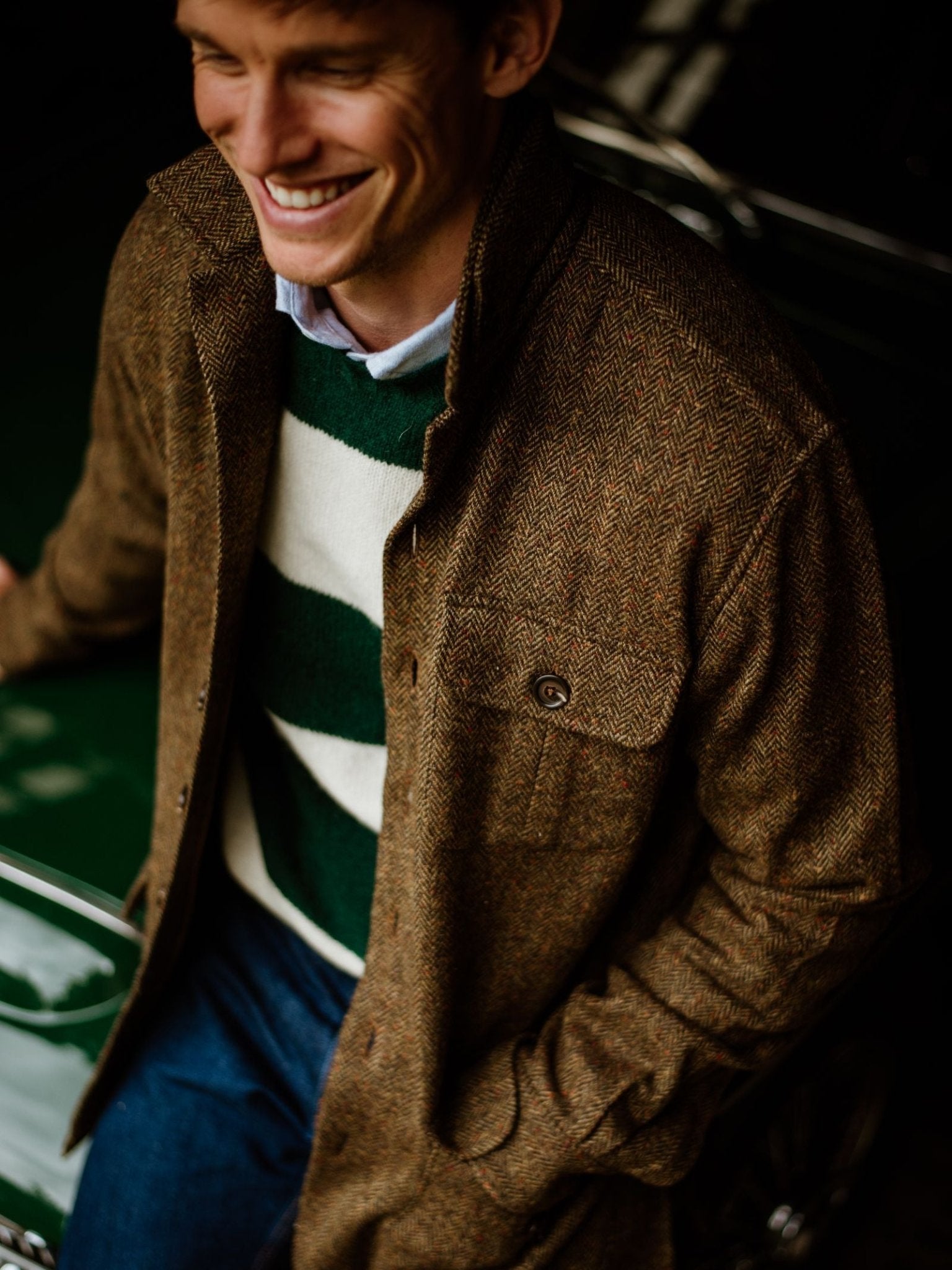 A smiling man in Campbell's of Beauly Casual Tweed Shacket, layered over a green and white striped sweater and blue jeans, leans against a green car, embodying relaxed tailoring with effortless style.