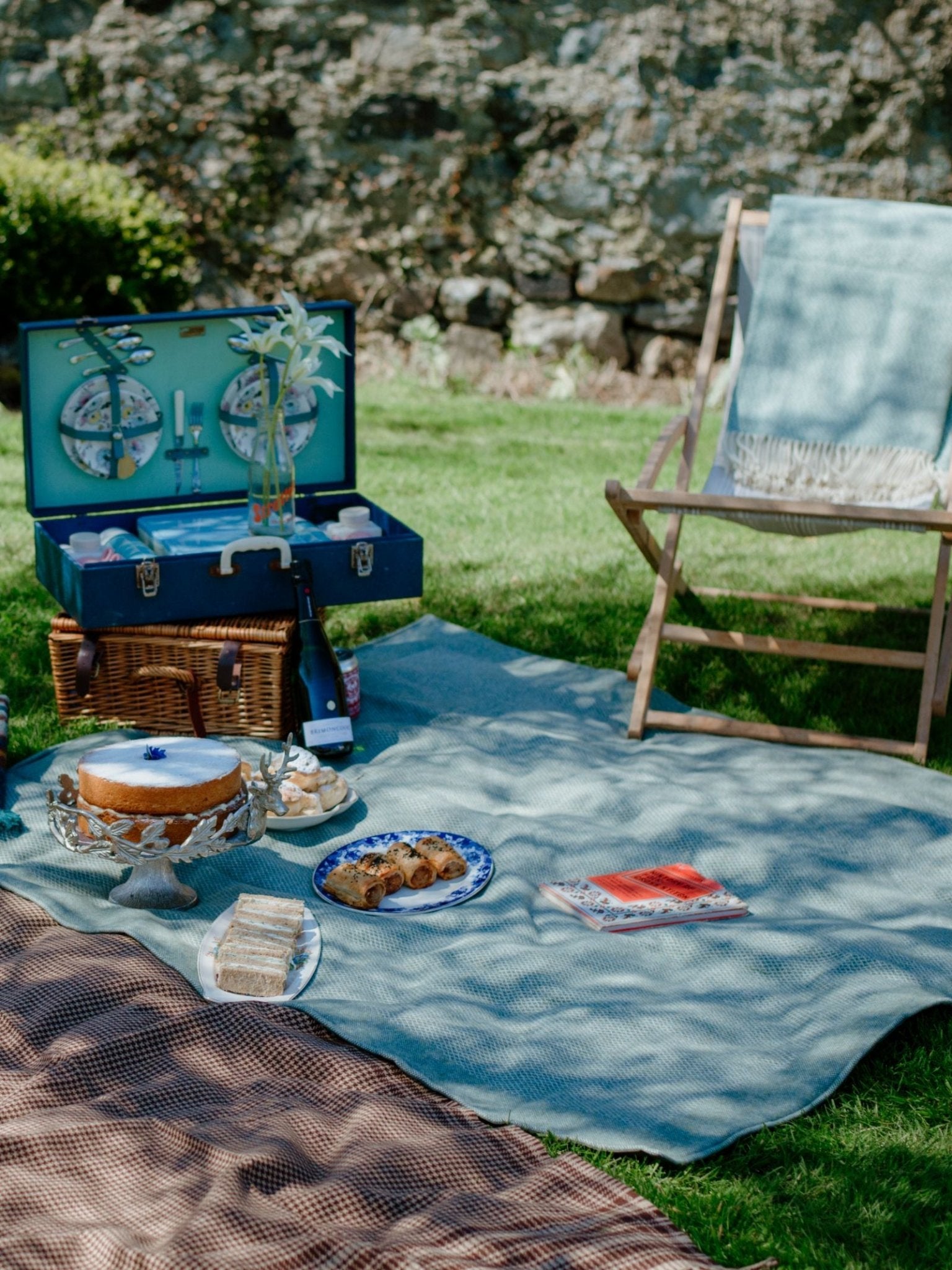 A picnic setup on grass features the Campbells of Beauly Waterproof Picnic Blanket in blue with heritage weaving, a wicker basket, assorted food, a blue cutlery case, and a foldable chair beside a stone wall under daylight.