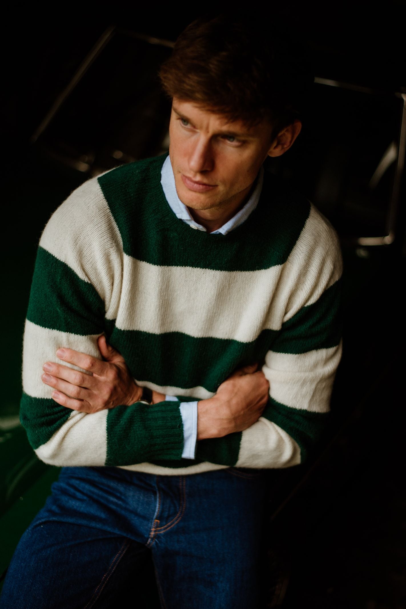 A man with short brown hair wears the Campbells of Beauly Shetland Stripe Crew Jumper and blue jeans, sitting with arms crossed and a serious expression. The dimly lit, blurred background keeps the focus on the stylish striped sweater.