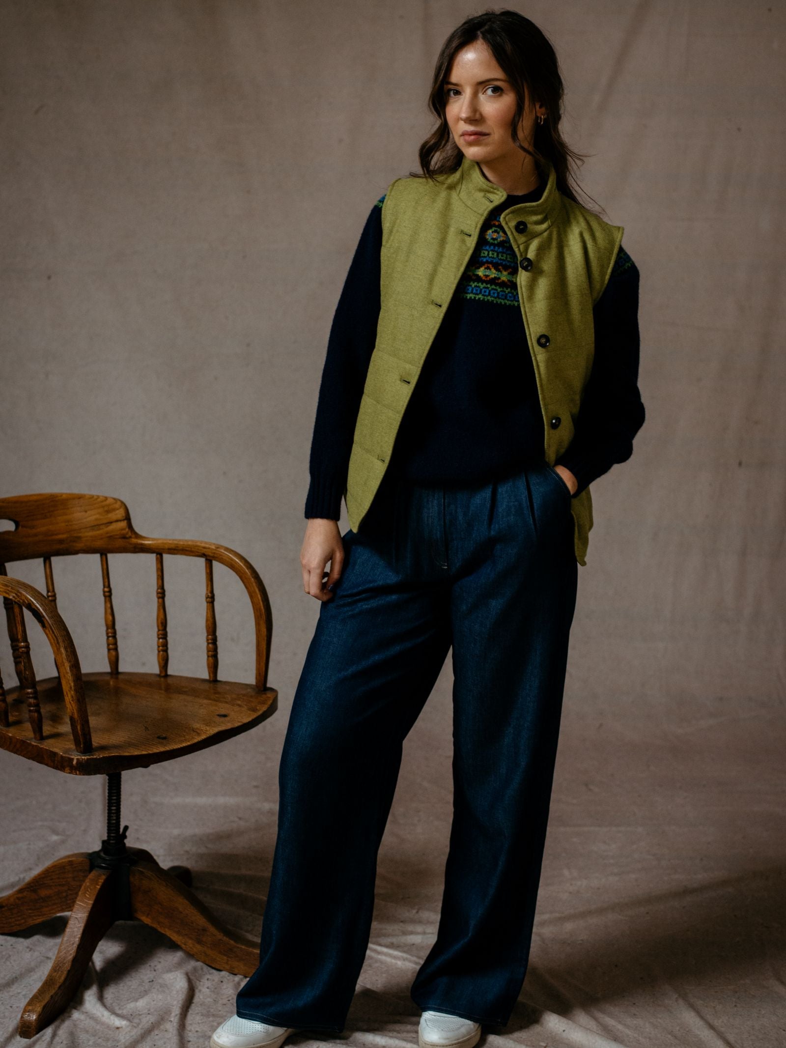 A woman with long brown hair stands indoors beside a wooden chair, wearing Campbell’s of Beaulys Quilted Buttoned Gilet over a dark patterned sweater, wide-leg blue pants, and white shoes. She has a neutral expression.