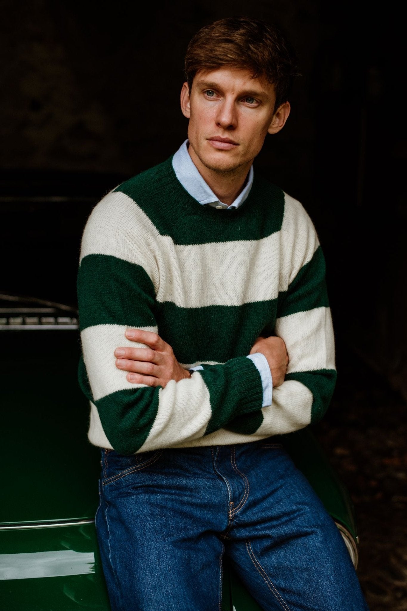 A young man with short brown hair wears a Campbells of Beauly Shetland Stripe Crew Jumper over a light blue collared shirt and blue jeans, sitting with arms crossed and gazing thoughtfully into the distance.