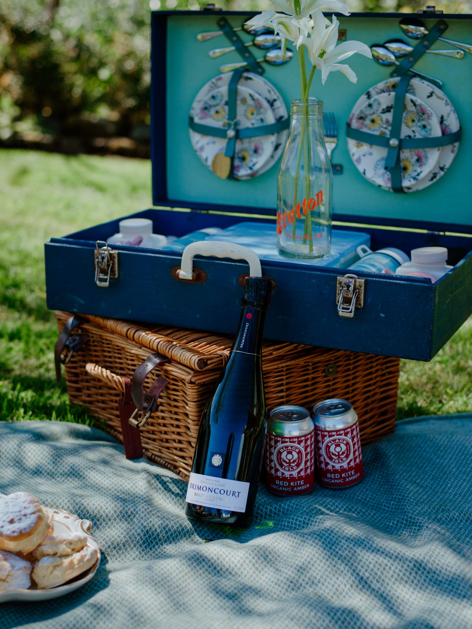 A picnic setup on a Campbells of Beauly Waterproof Picnic Blanket, featuring a wicker basket, Humoncourt sparkling wine, three Red Kite beers, scones, and a blue picnic box with plates, utensils, and white flowers in a vase.