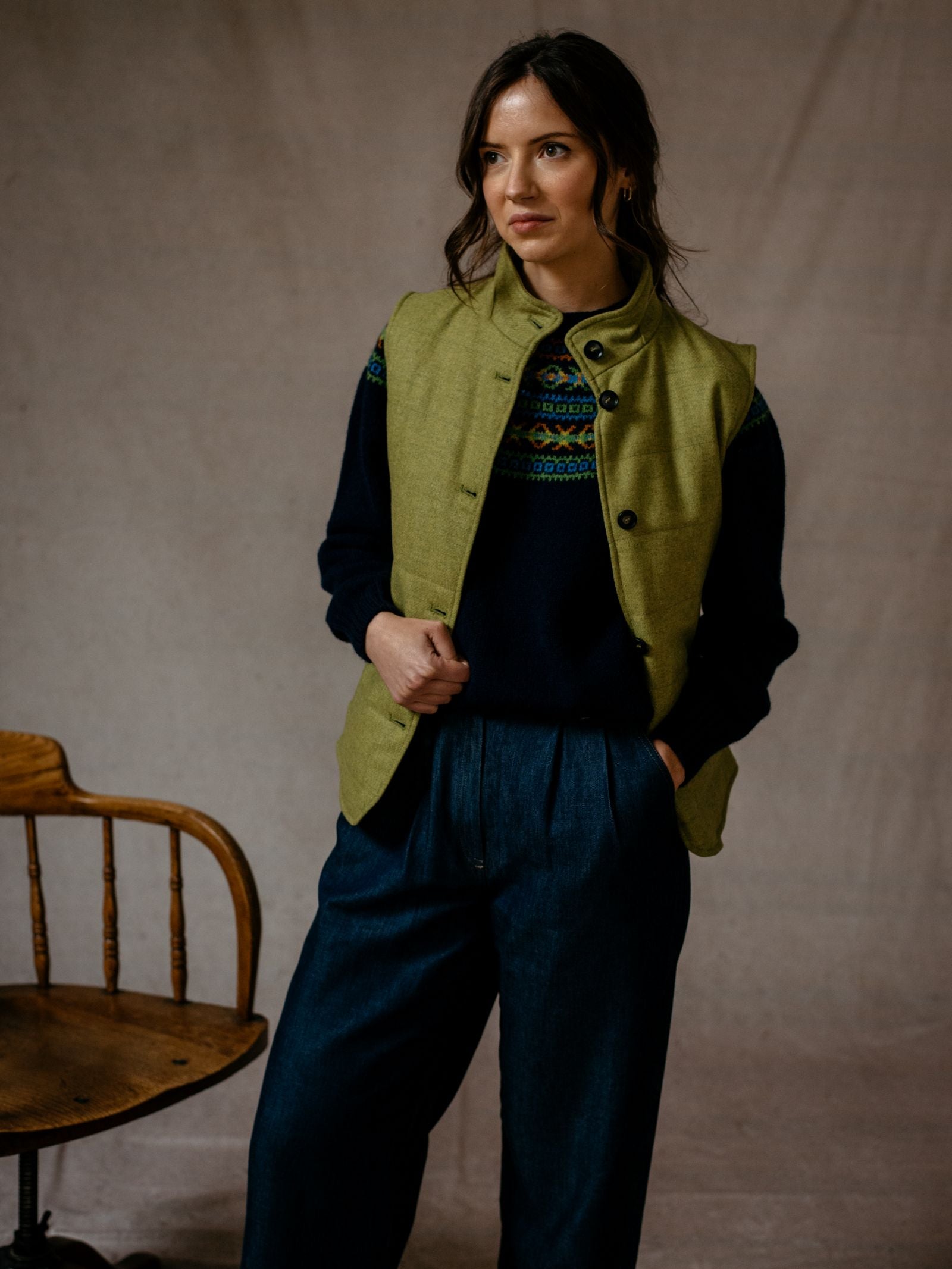 A woman stands indoors with her hands in her pockets, wearing the Campbells of Beauly Quilted Buttoned Gilet over a patterned navy sweater and wide-leg dark trousers, posing beside a wooden chair.