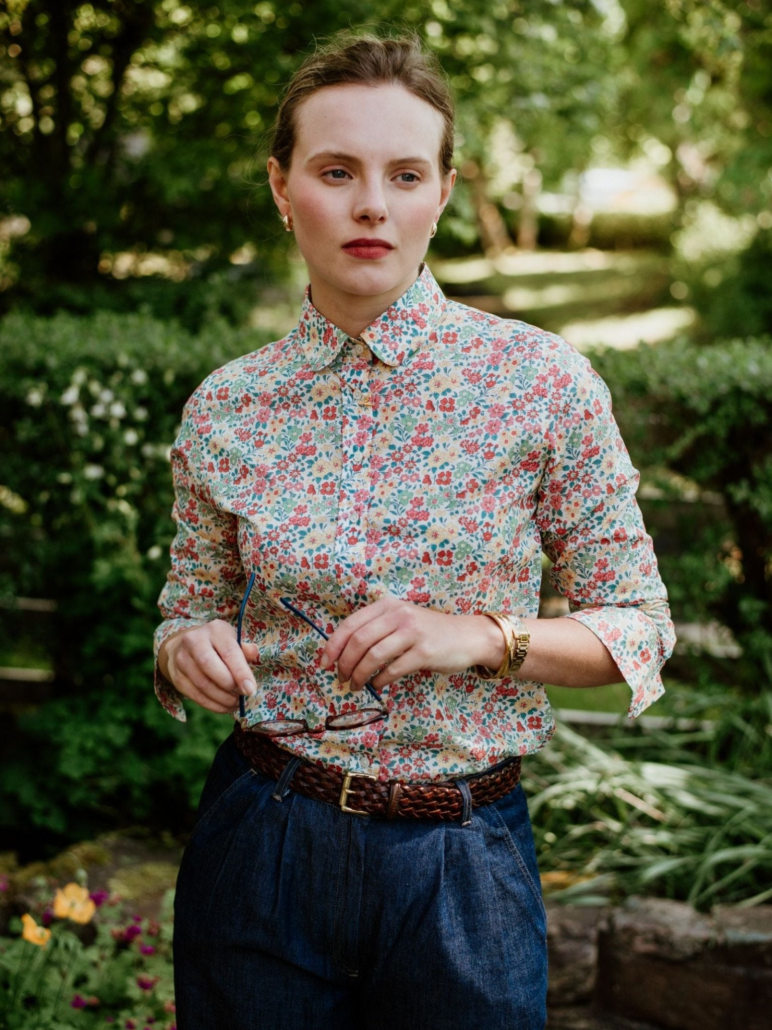 A woman stands outdoors amid lush greenery, wearing a Campbells of Beauly Liberty Print Cotton Shirt tucked into high-waisted blue pants with a braided brown belt, holding eyeglasses in one hand.