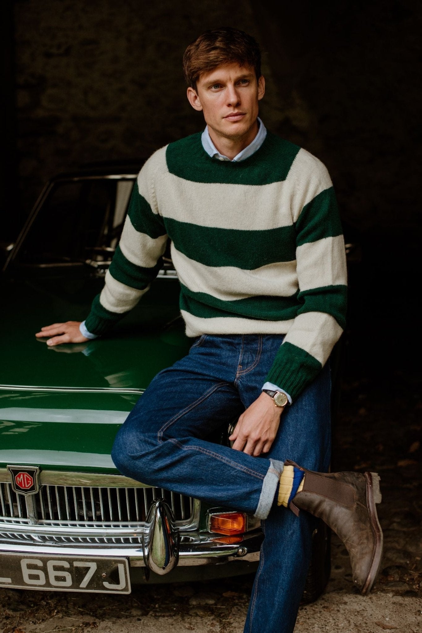 A man in the Campbells of Beauly Shetland Stripe Crew Jumper sits on a classic green MG car, pairing the green and white striped sweater with blue jeans and brown boots, gazing thoughtfully to the side.