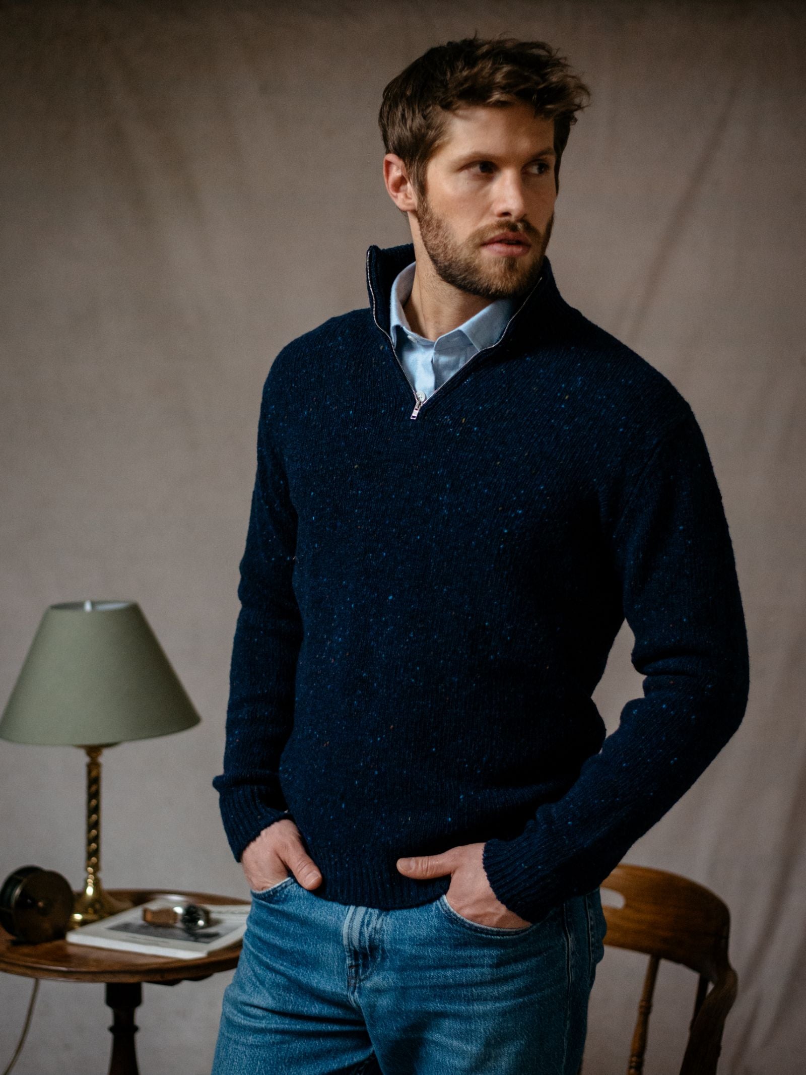 A man with short brown hair and a beard wears the Campbells of Beauly Donegal Lambswool Zip Neck Jumper in navy over a light blue shirt, hands in jean pockets. In the background, a table holds books, glasses, and a green lamp.