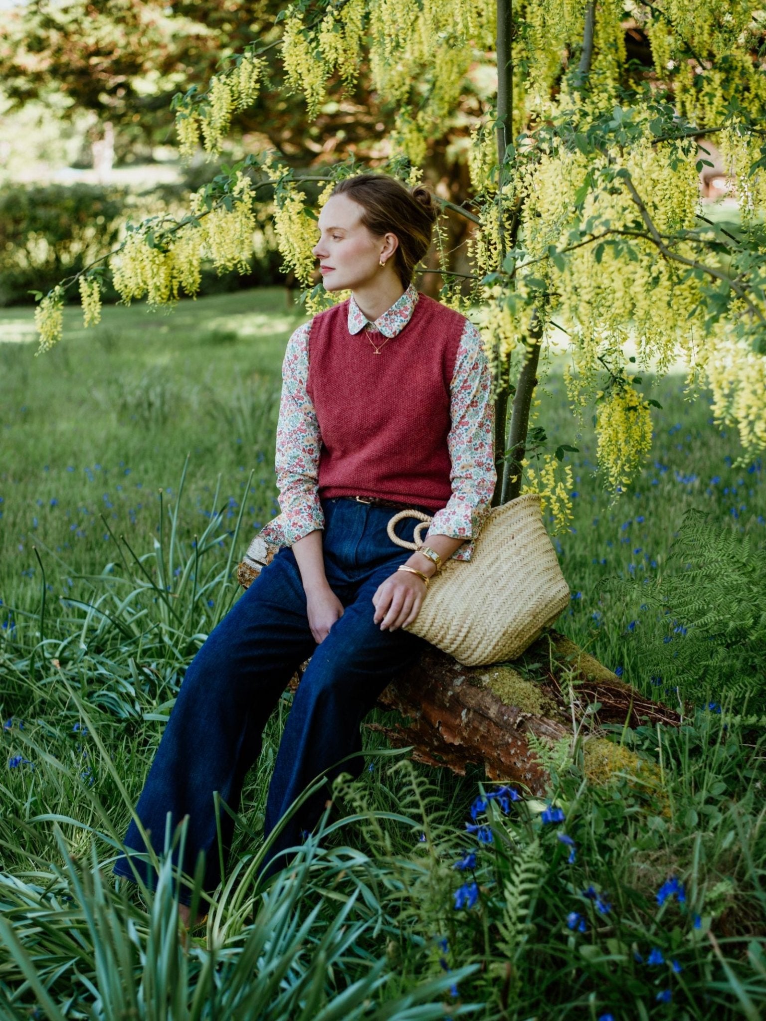 A woman in a red sweater vest and Campbells of Beauly Liberty Print Cotton Shirt sits on a fallen log in a grassy garden, surrounded by green plants and yellow flowers, holding a woven straw bag.