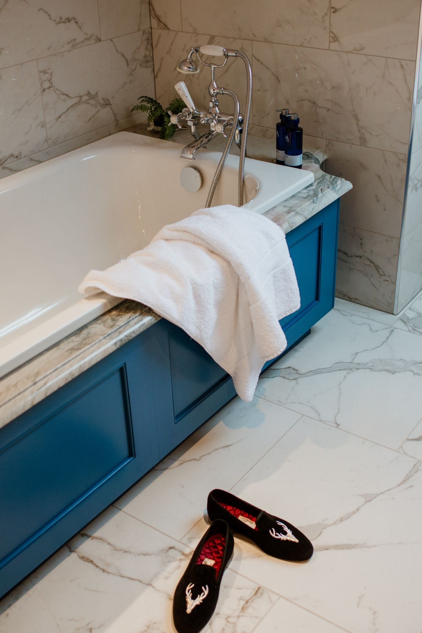A white towel hangs over a blue-paneled bathtub in a marble-tiled bathroom. Campbells of Beauly Stag Velvet Slippers are on the floor nearby, with toiletries and a small plant on the tub’s ledge.