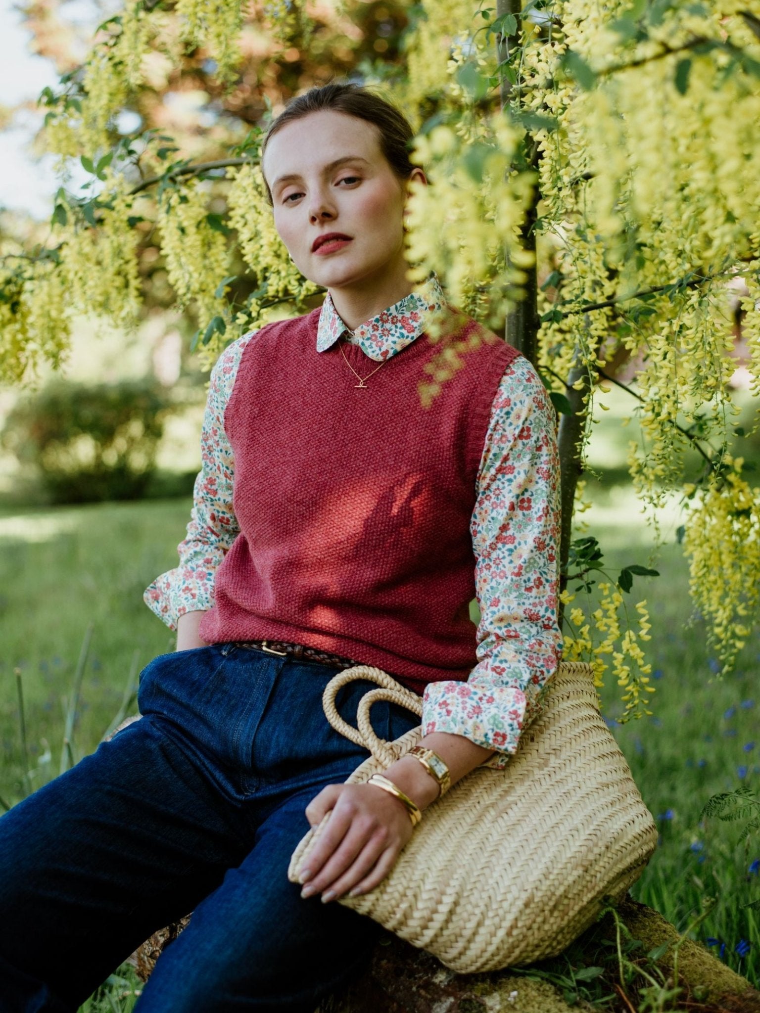 A woman sits outdoors on a rock under a tree, wearing blue jeans and the Campbells of Beauly Moss Stitch Crew Tank layered over her floral shirt. With her woven straw bag, she looks calmly at the camera, showcasing versatile spring style.
