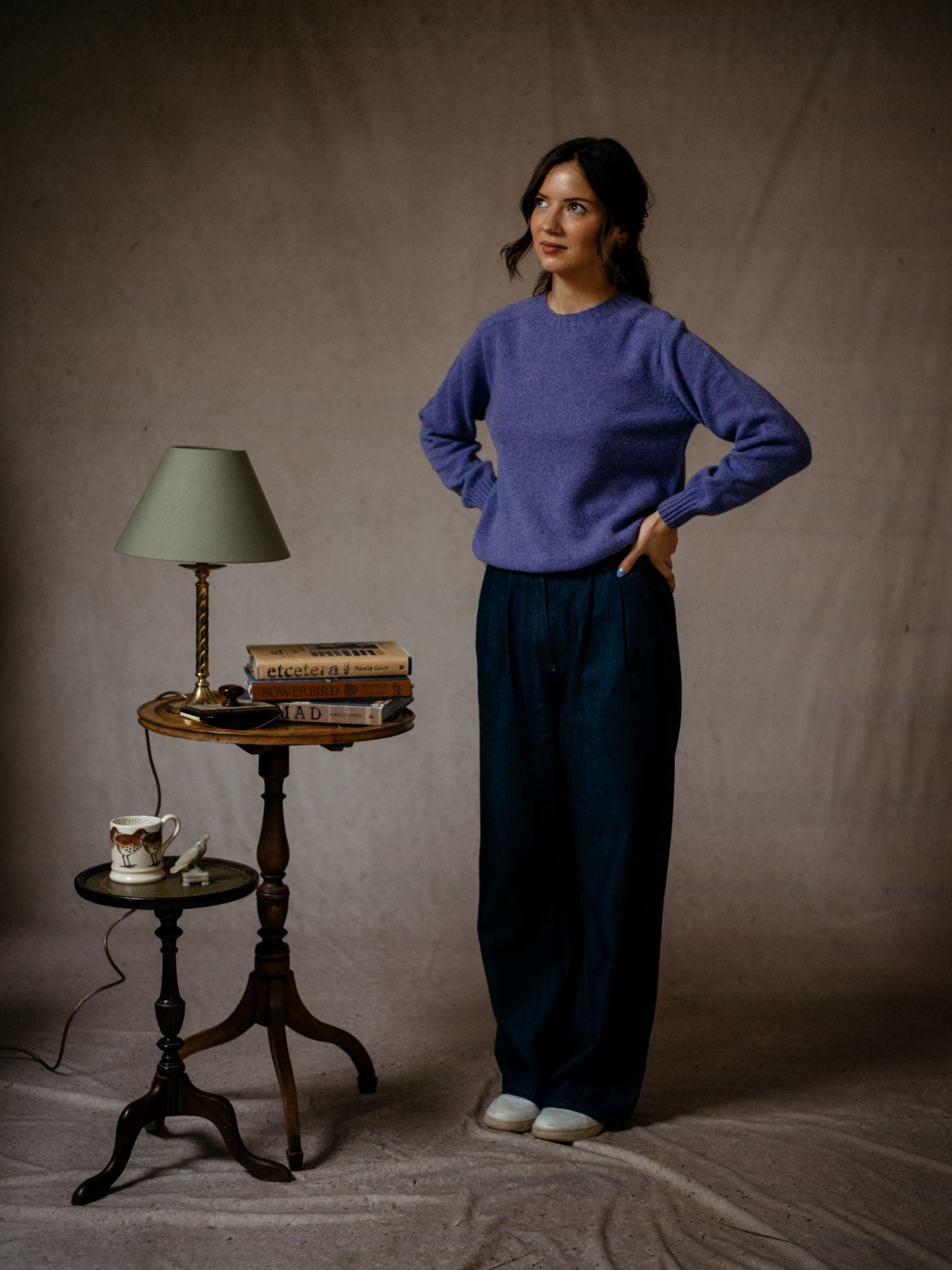 A woman wearing a purple Geelong Lambswool Crew Neck Jumper by Campbells of Beauly and wide-leg pants stands with hands on hips beside a small table with a green lamp, books, and teacup against a neutral fabric backdrop.