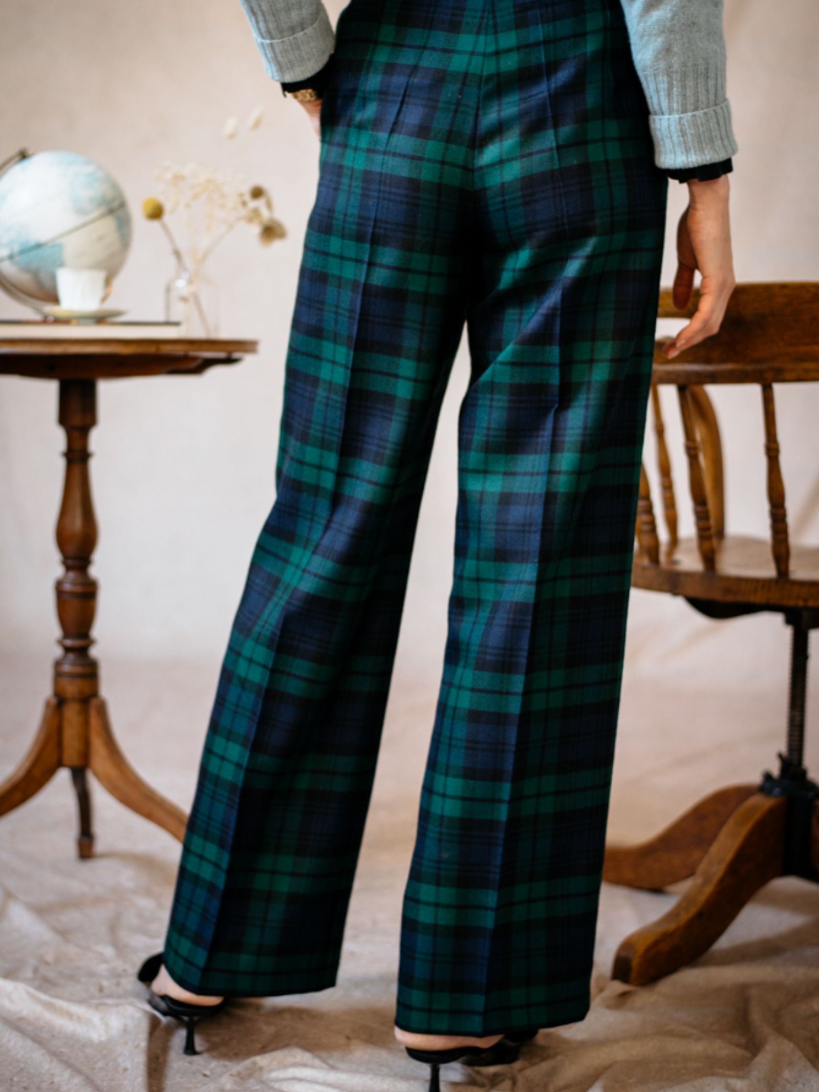 A person stands indoors wearing Campbells of Beauly Wide Leg Blackwatch Trousers with black heels. Part of their upper body is visible. In the background, a small wooden table displays a globe and decorative items.