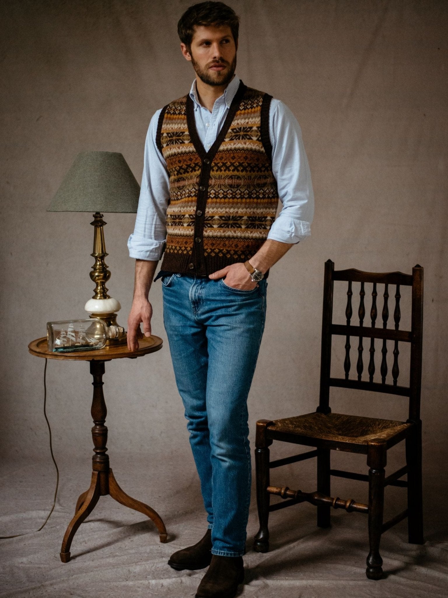 A man in a Campbells of Beauly Fairisle V-Neck Waistcoat, light blue shirt, and jeans stands next to a vintage wooden chair and round side table with a lamp, book, and glass case against a plain backdrop.