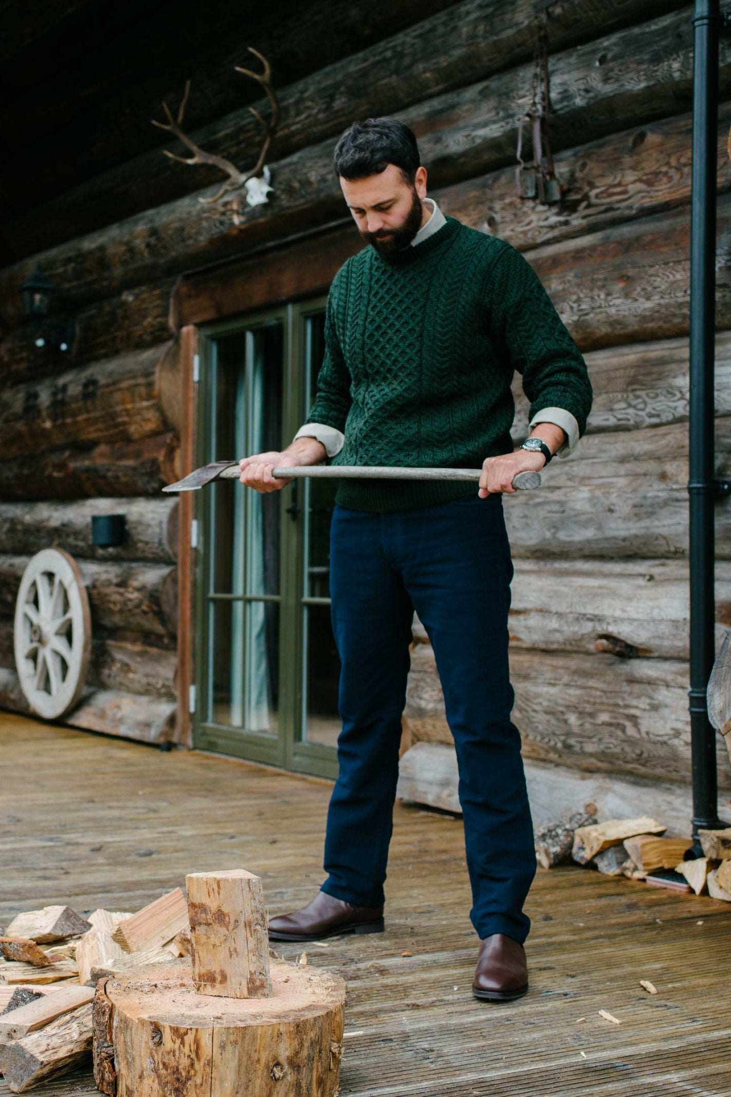 A man in a green sweater and dark pants—Campbells of Beauly Moleskin Jeans—stands on a wooden deck by a rustic log cabin, holding an axe as he prepares to chop firewood amid scattered logs.