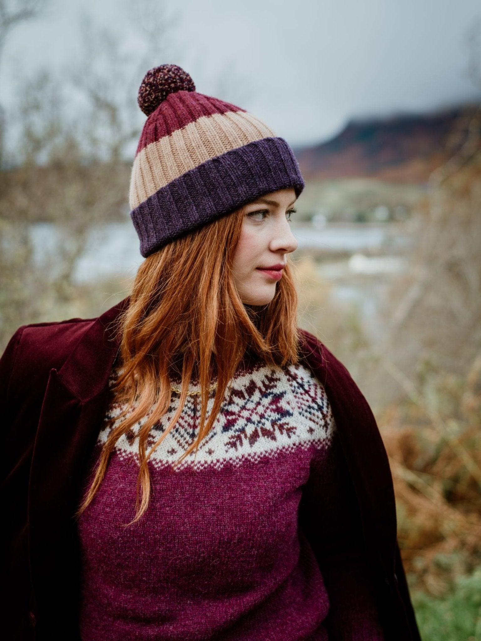 A woman with long red hair stands outside in the Scottish Borders, wearing the Campbells of Beauly Lambswool Stripe Hat with Pom-Pom and a patterned sweater. Blurred trees and mountains are visible in the background.