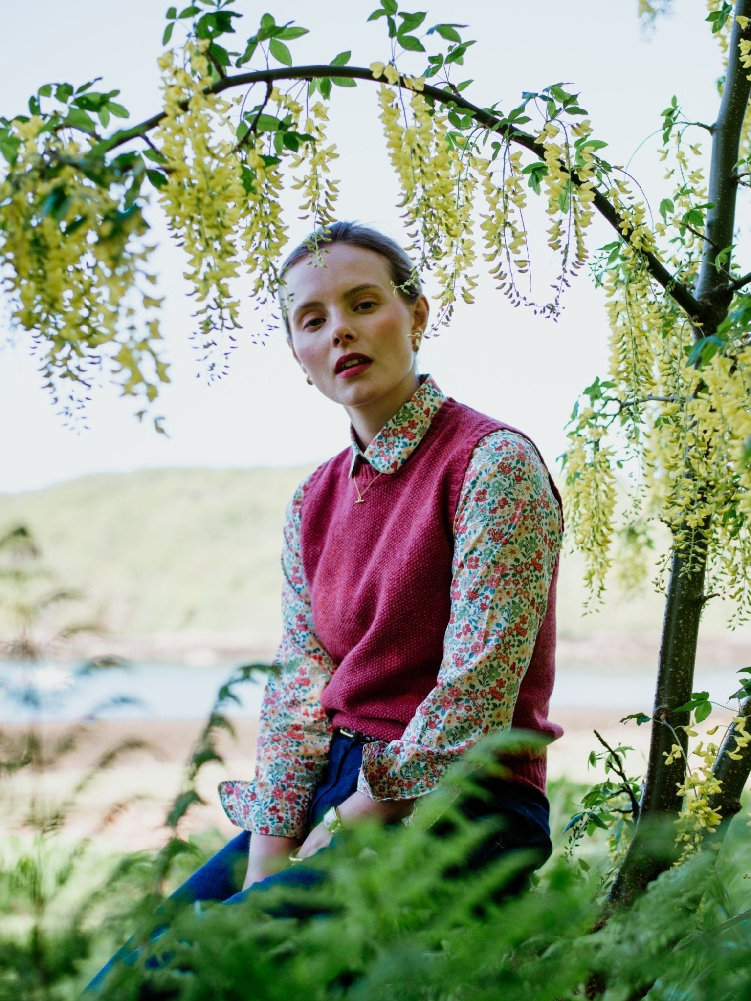 A person wearing a floral shirt and the Campbells of Beauly Moss Stitch Crew Tank sits outdoors beneath a tree with yellow flowers, surrounded by green foliage, with blurred hills and water in the background.