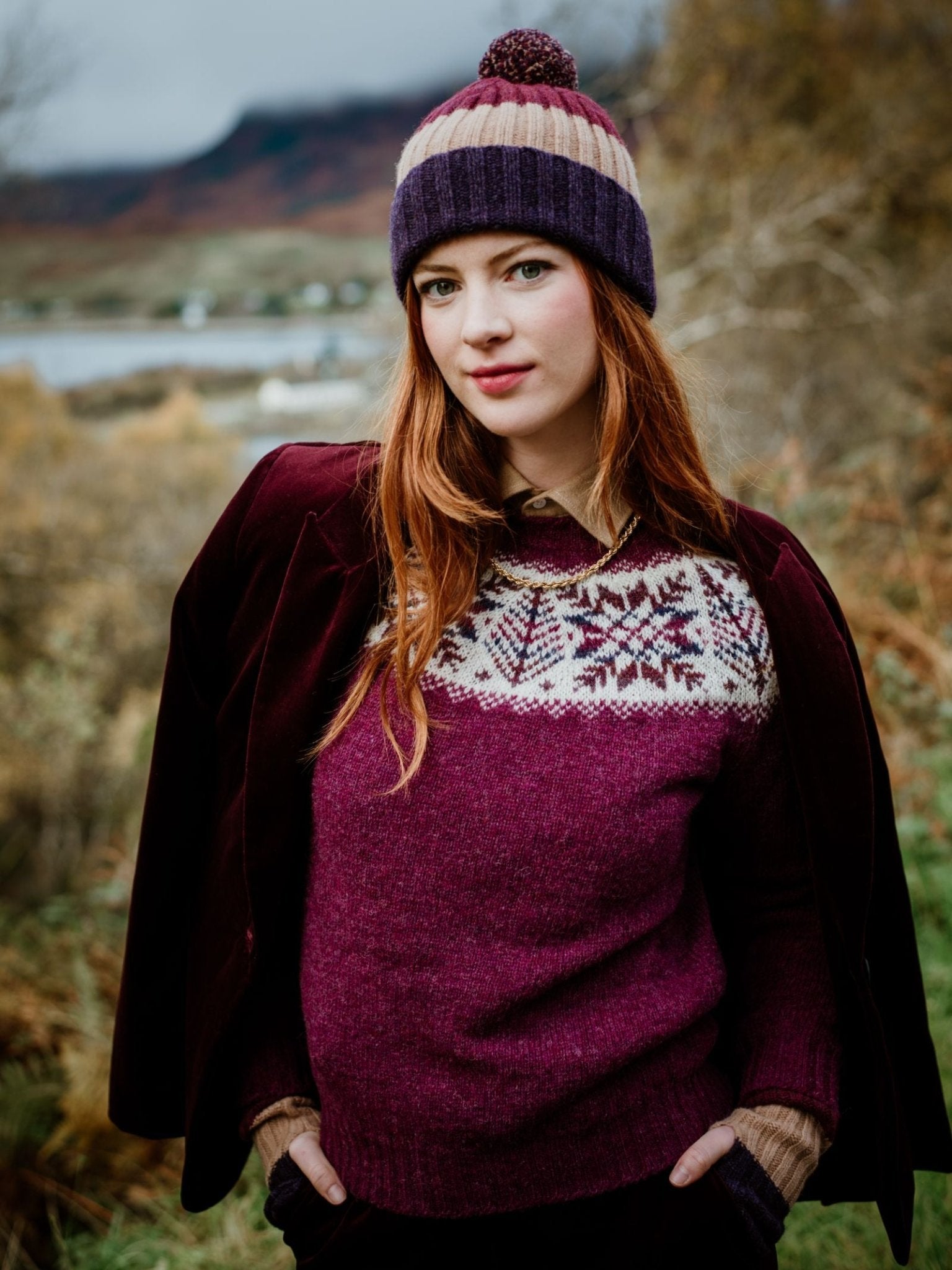 A woman with long red hair stands outdoors in the Scottish Borders, wearing the Campbells of Beauly Lambswool Stripe Hat with Pom-Pom. She has a patterned sweater and maroon coat draped over her shoulders, set against a scenic, blurred background.