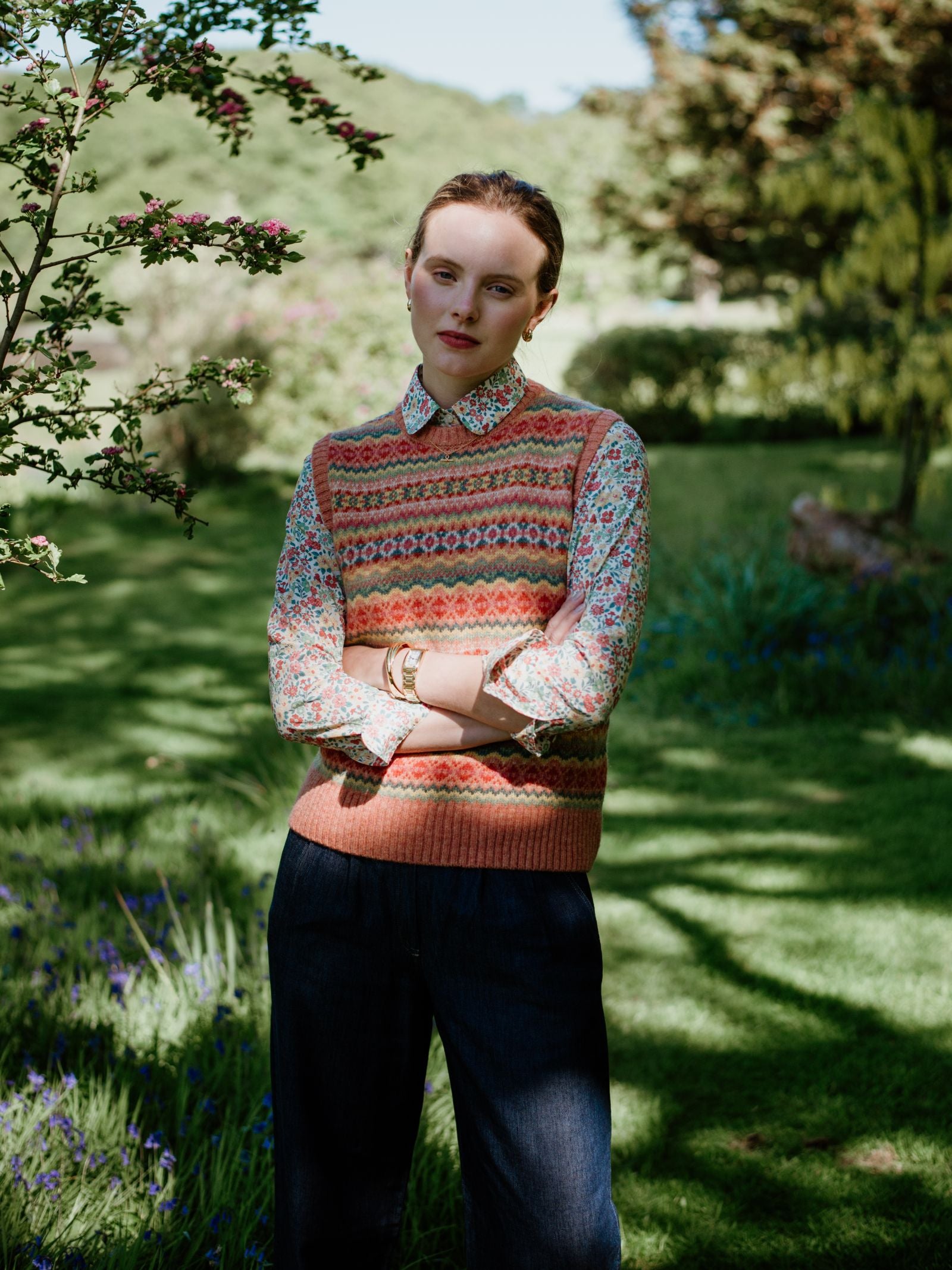 Outdoors on green grass, a person wears dark pants and a Campbells of Beauly Fairisle Crew Tank with a traditional pattern over a floral shirt, accessorized with gold bracelets. Their arms are crossed amid sunlit trees.