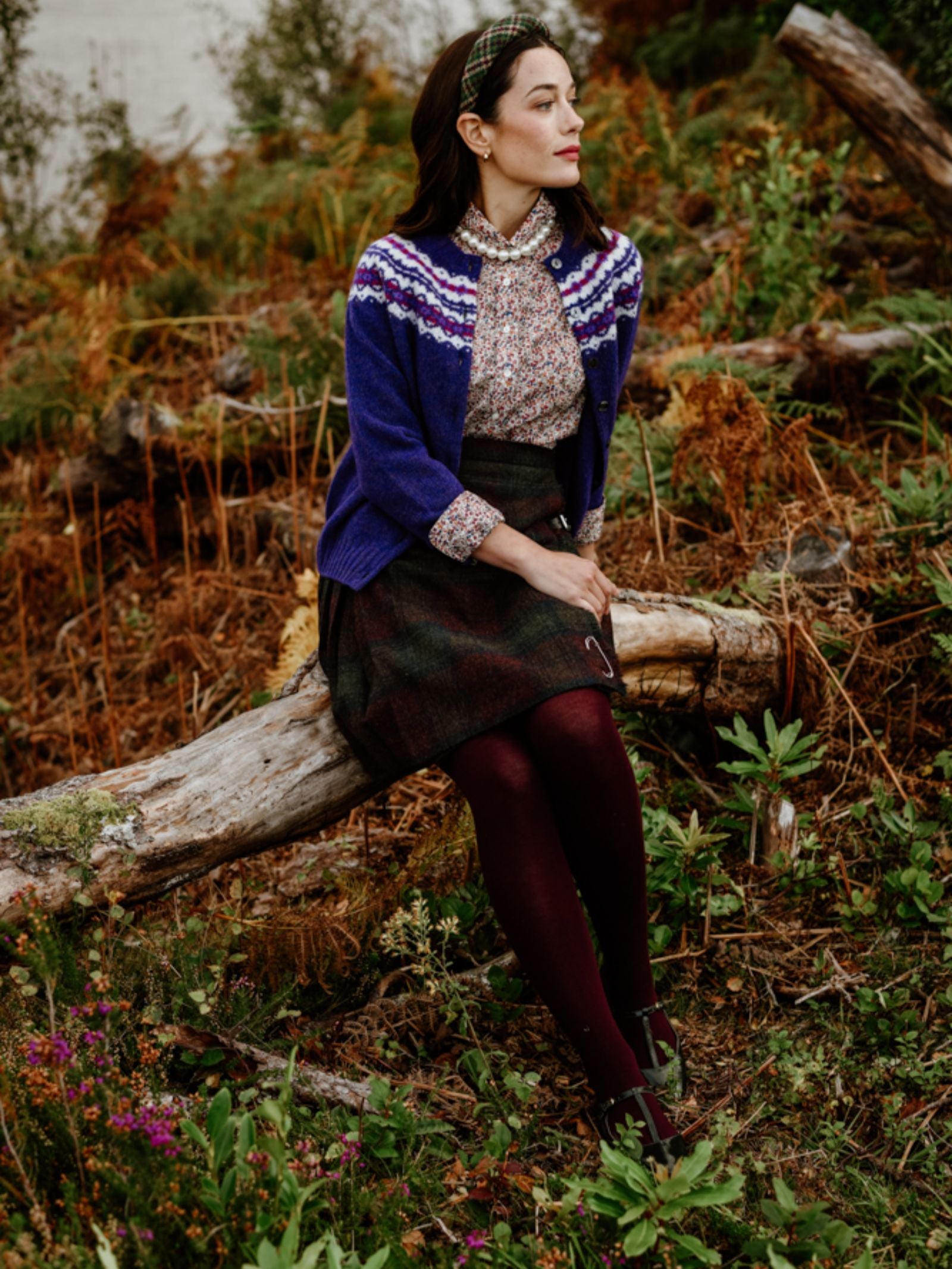 A woman in a Campbell's of Beauly Shetland Fairisle Cardigan, floral blouse, and dark skirt sits thoughtfully on a fallen log in a lush green outdoor setting.