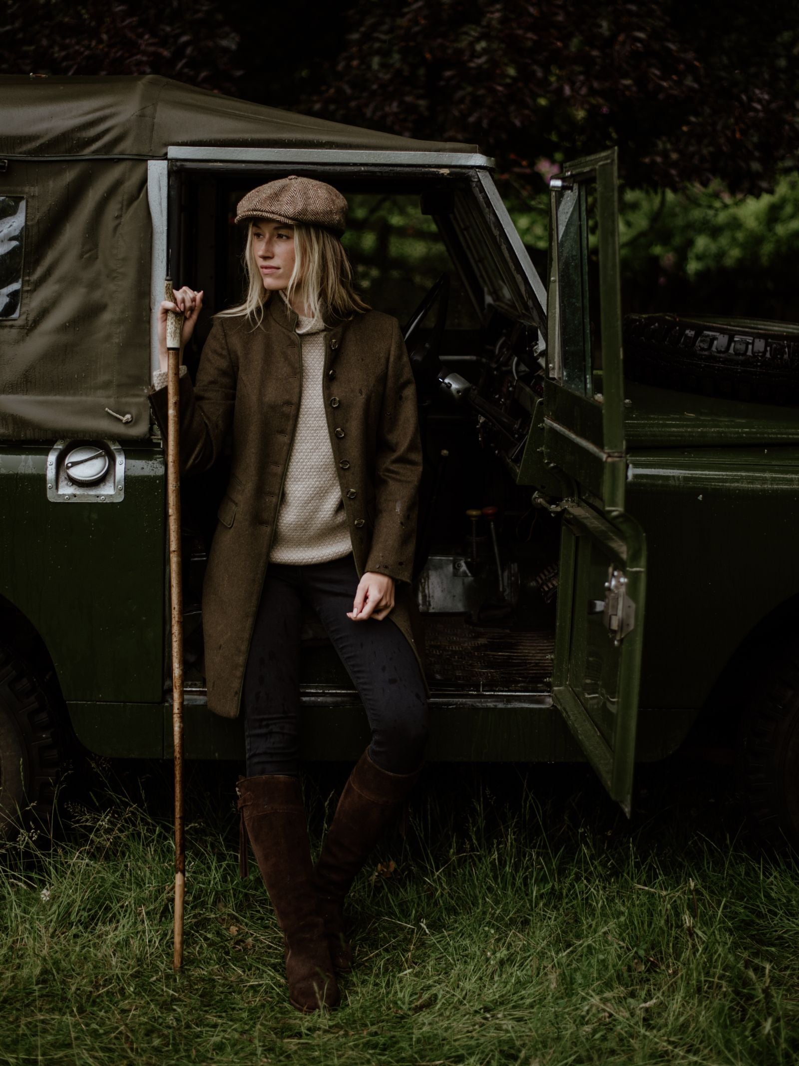 A person in a flat cap and coat, wearing Campbells of Beauly Suede Spanish Boots, leans on a wooden stick beside an open door of a classic green off-road vehicle, standing on grass with trees behind.