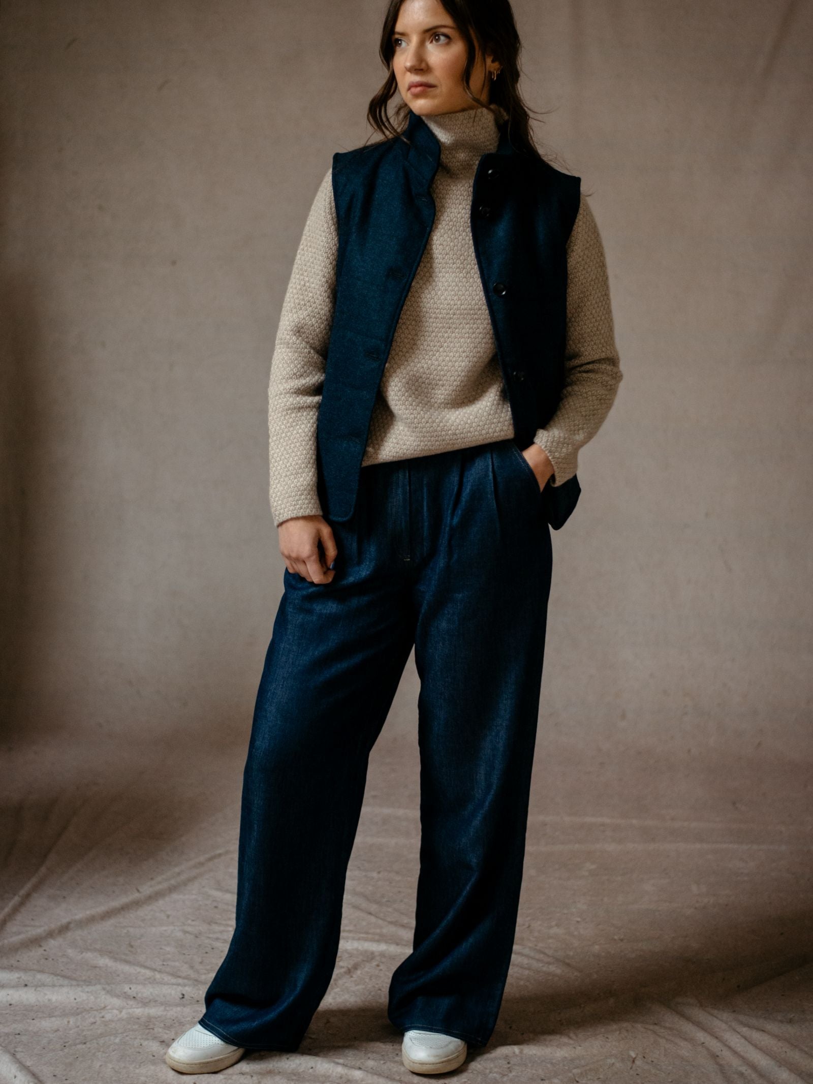 A woman stands against a neutral backdrop wearing a cream turtleneck, the Campbells of Beauly Quilted Buttoned Gilet in navy tweed, loose navy trousers, and white shoes, with her left hand in her pocket.