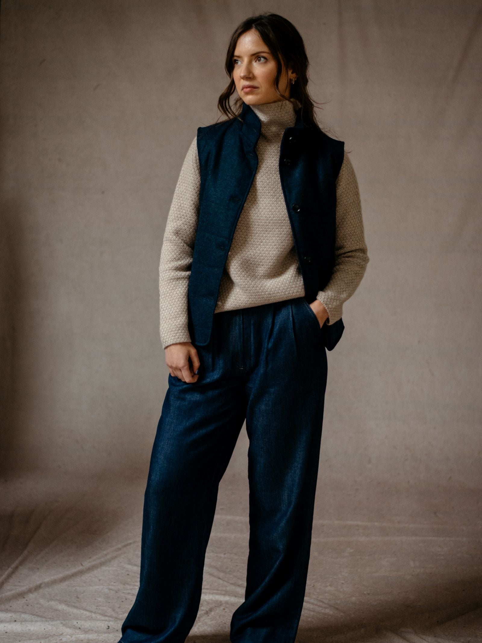 A woman stands against a neutral background wearing Campbells of Beauly Wide Leg Jeans, styled with a cream turtleneck sweater and dark blue vest. She has long brown hair and gazes thoughtfully to the side.