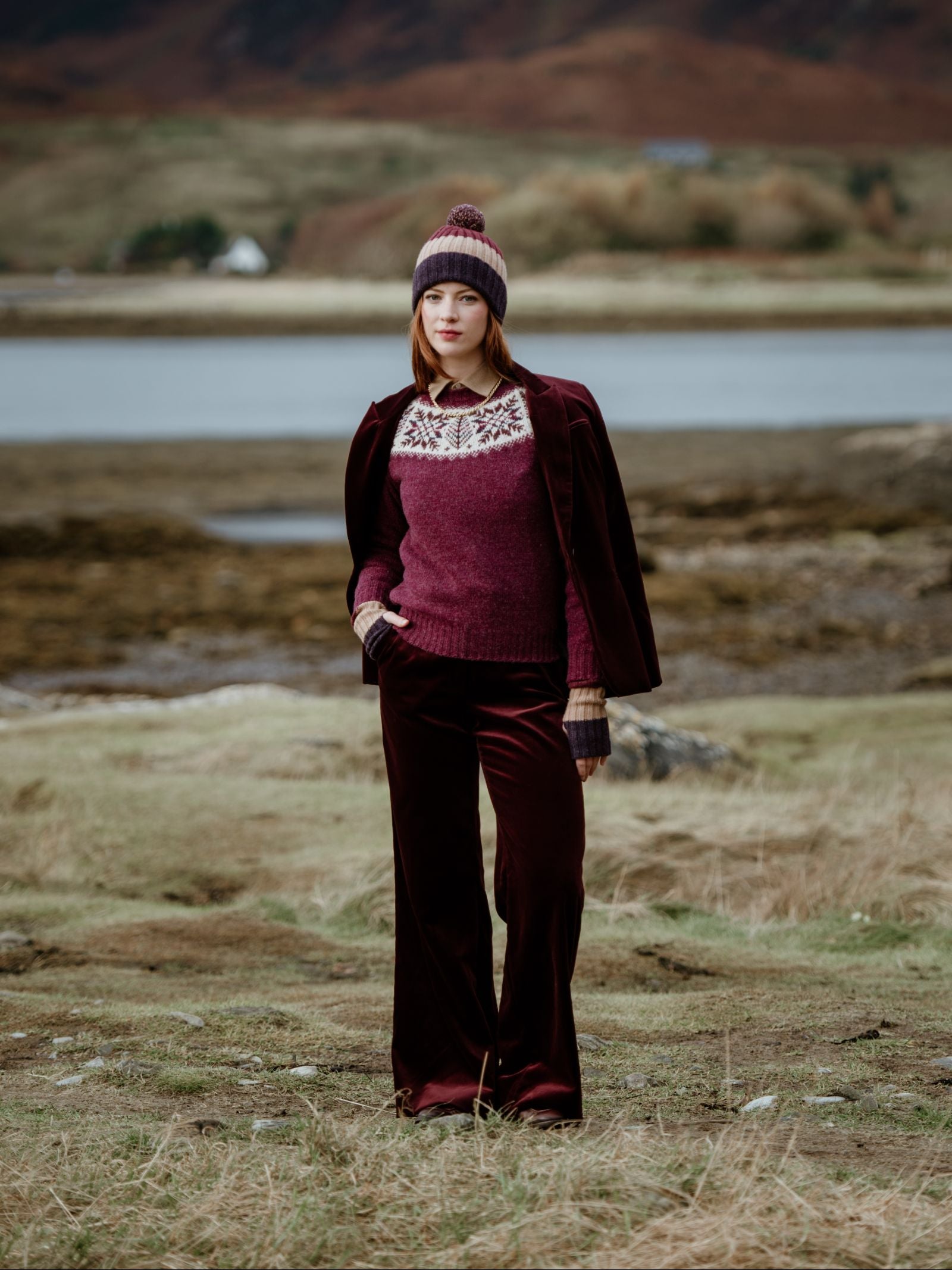 A person stands outdoors in the Scottish Borders, wearing a maroon velvet suit, a patterned sweater, knit gloves, and the Campbells of Beauly Lambswool Stripe Hat with Pom-Pom. Grassy hills, houses, and trees are visible in the background.