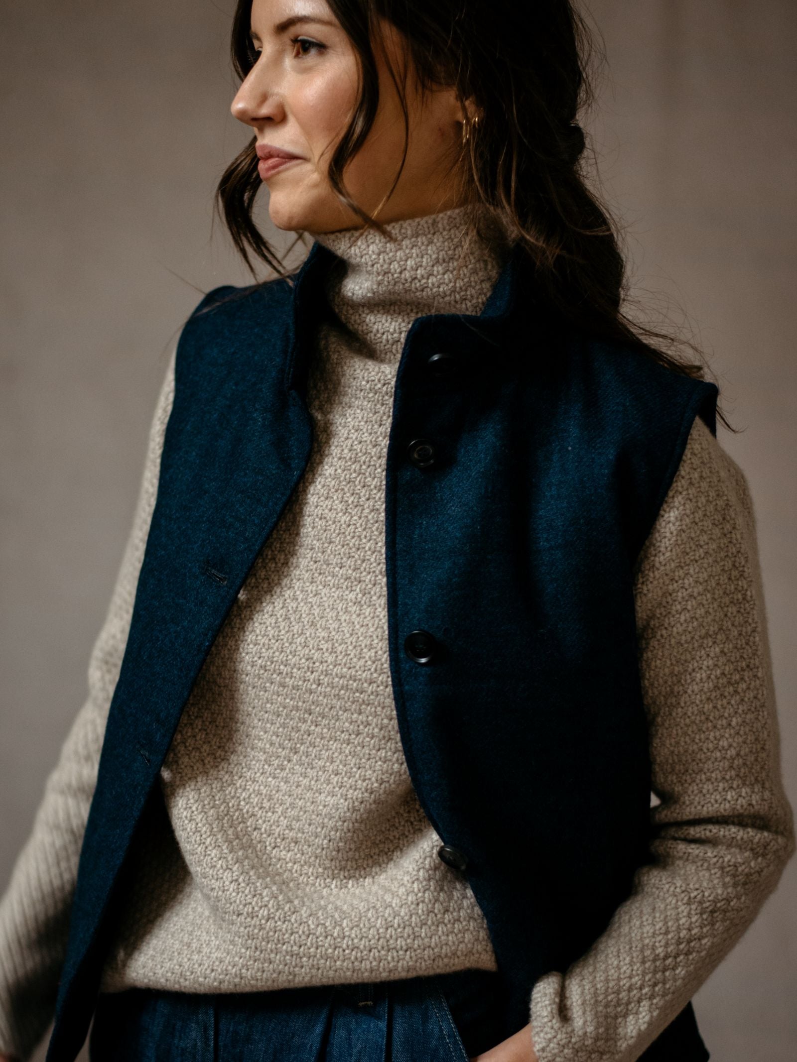 A woman with wavy brown hair wears the Campbells of Beauly Quilted Buttoned Gilet—a versatile layering piece inspired by British tweed—over a beige turtleneck, standing against a neutral background and gazing gently to the side.