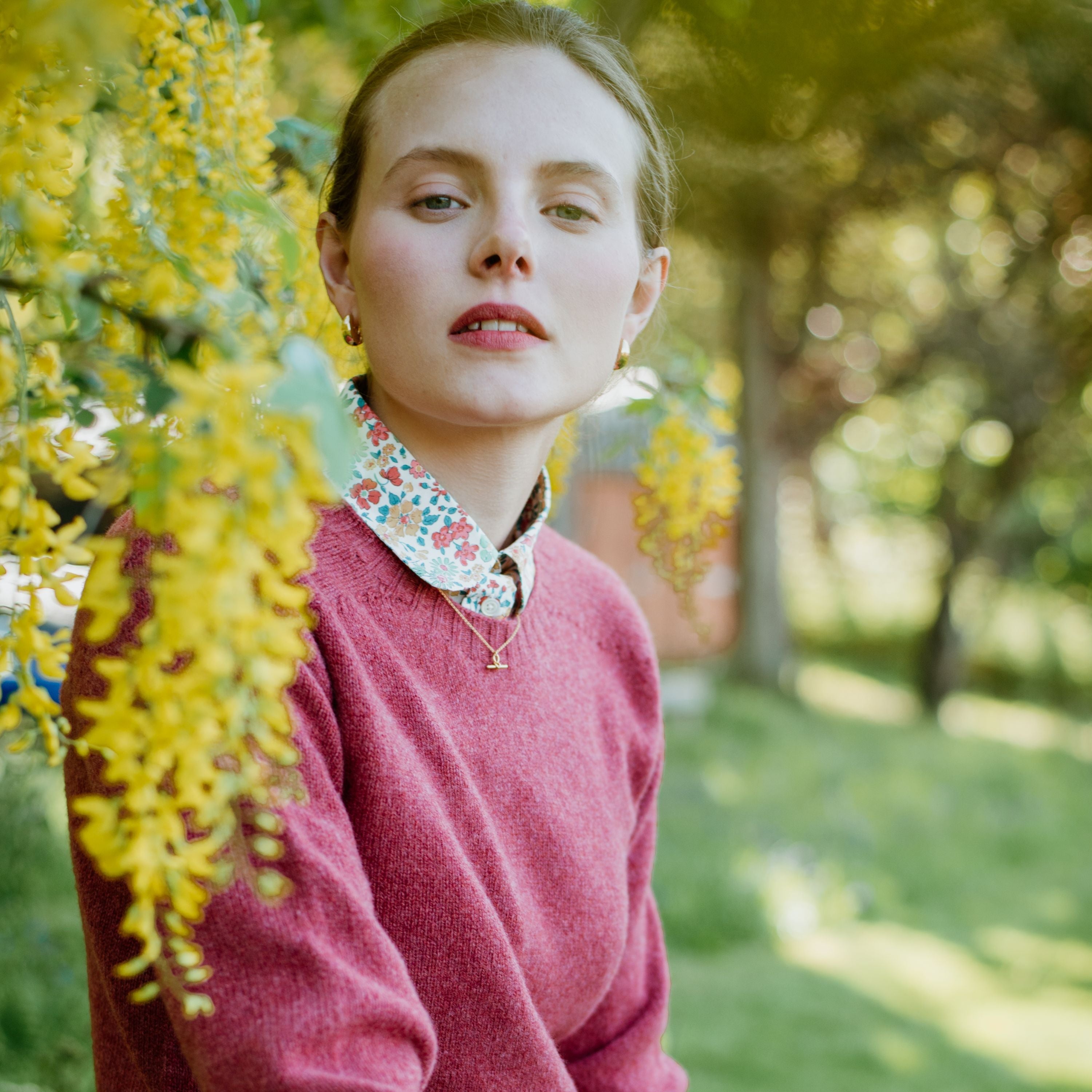 A young woman wearing a red sweater and floral collared shirt sits outdoors beside a branch with hanging yellow flowers, with greenery and sunlight in the blurred background.